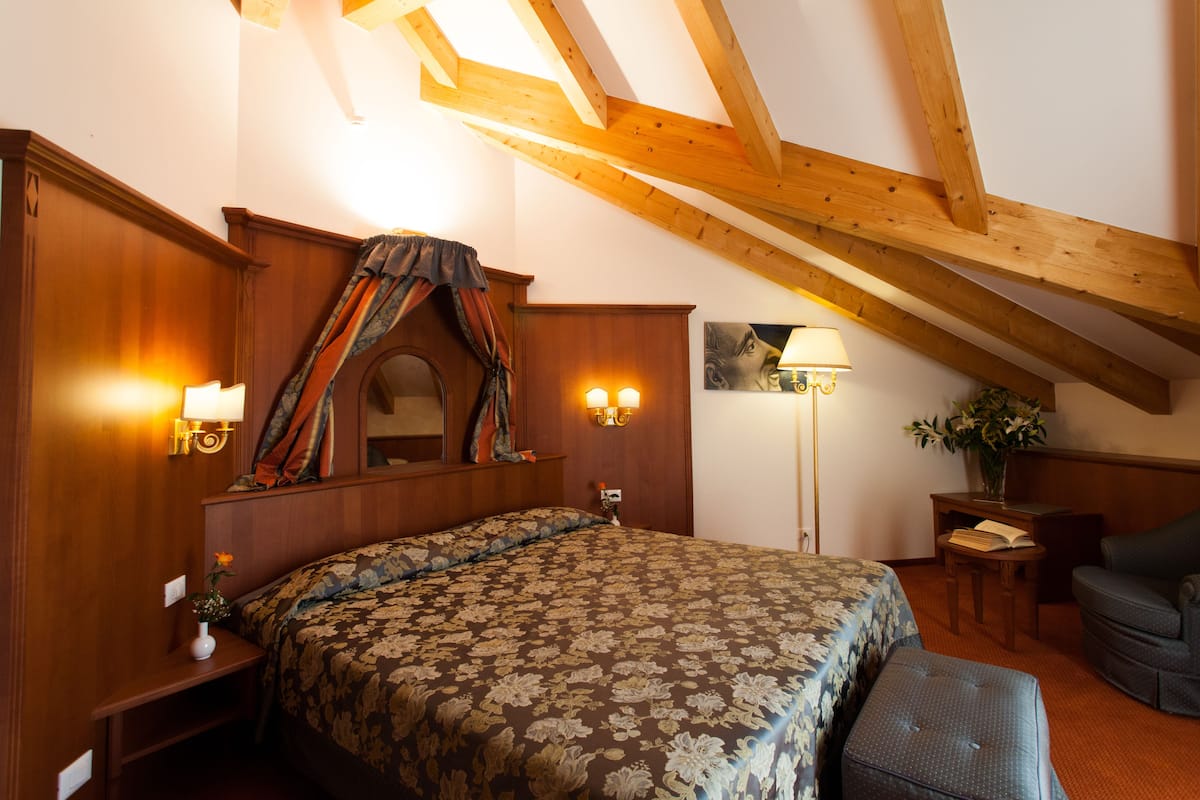 A bedroom with a wooden ceiling, a bed with a floral bedspread, a nightstand with a lamp, and a chair.