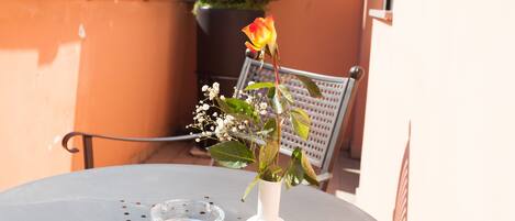 Una mesa con un jarrón de flores y un vaso de agua en un balcón, con una silla y una planta en maceta al fondo.