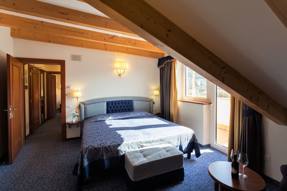 A bedroom with a large bed, bedside lamps, a small table with a bottle and glass, and a window with curtains.