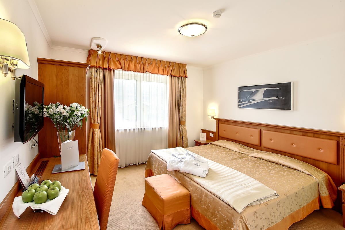 A hotel room with a bed, a desk with a lamp, a chair, a TV, and a painting on the wall.