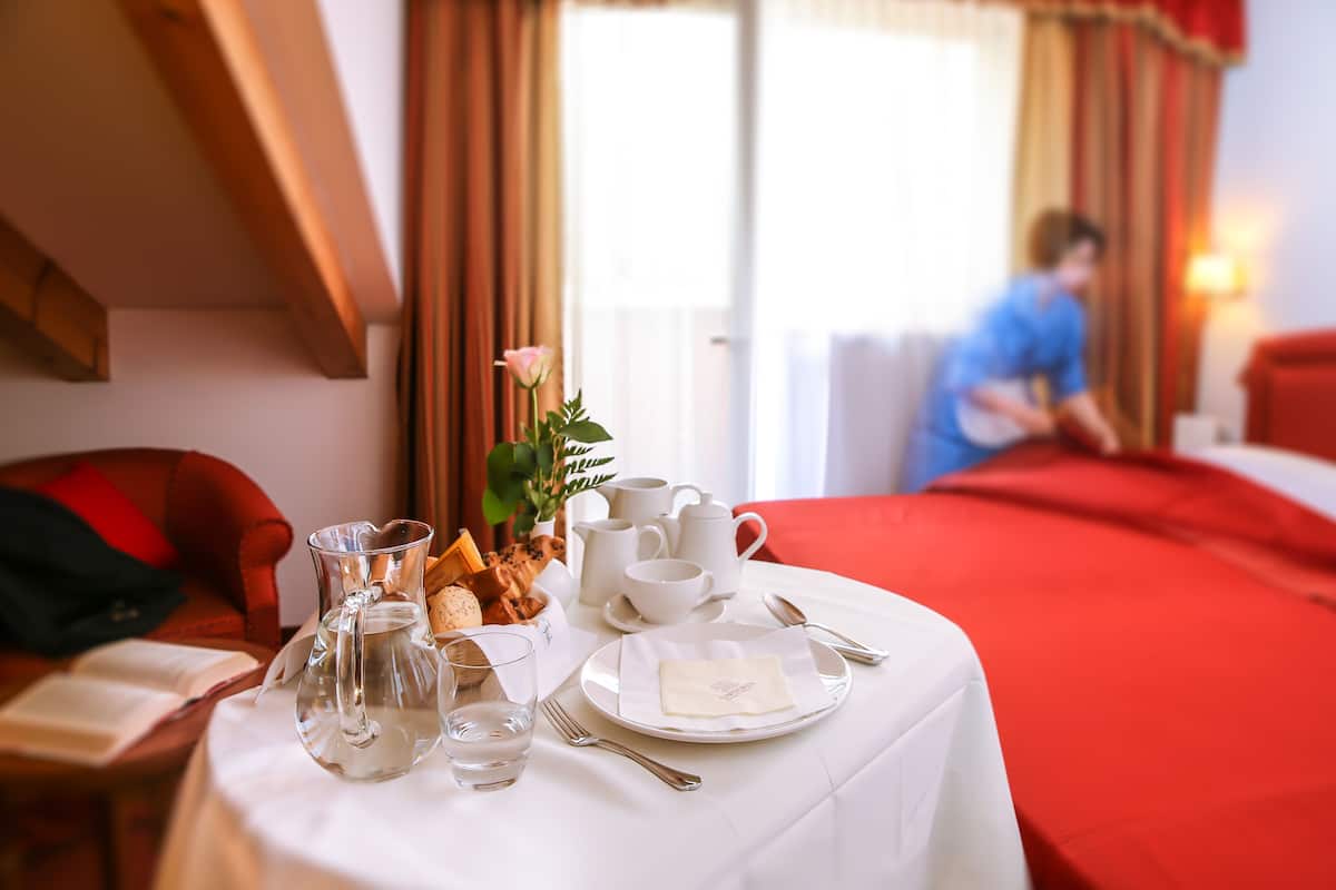 A neatly set dining table with a pitcher, cups, and a plate of food, in a room with a red bedspread and curtains.