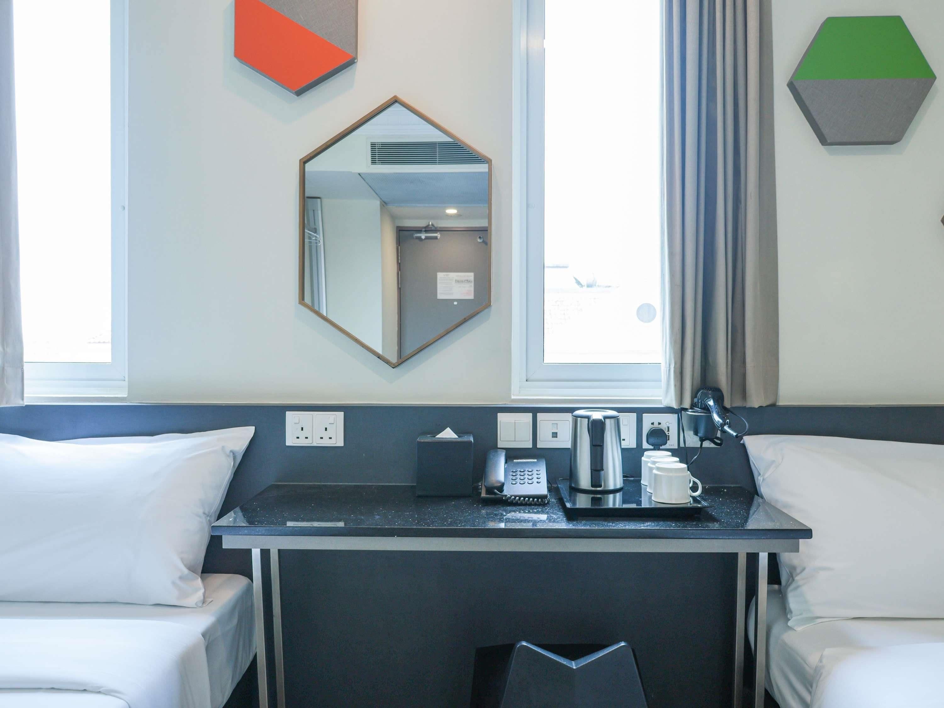 A hotel room with a desk, two white beds, a mirror, a window, and a green and gray wall decoration.