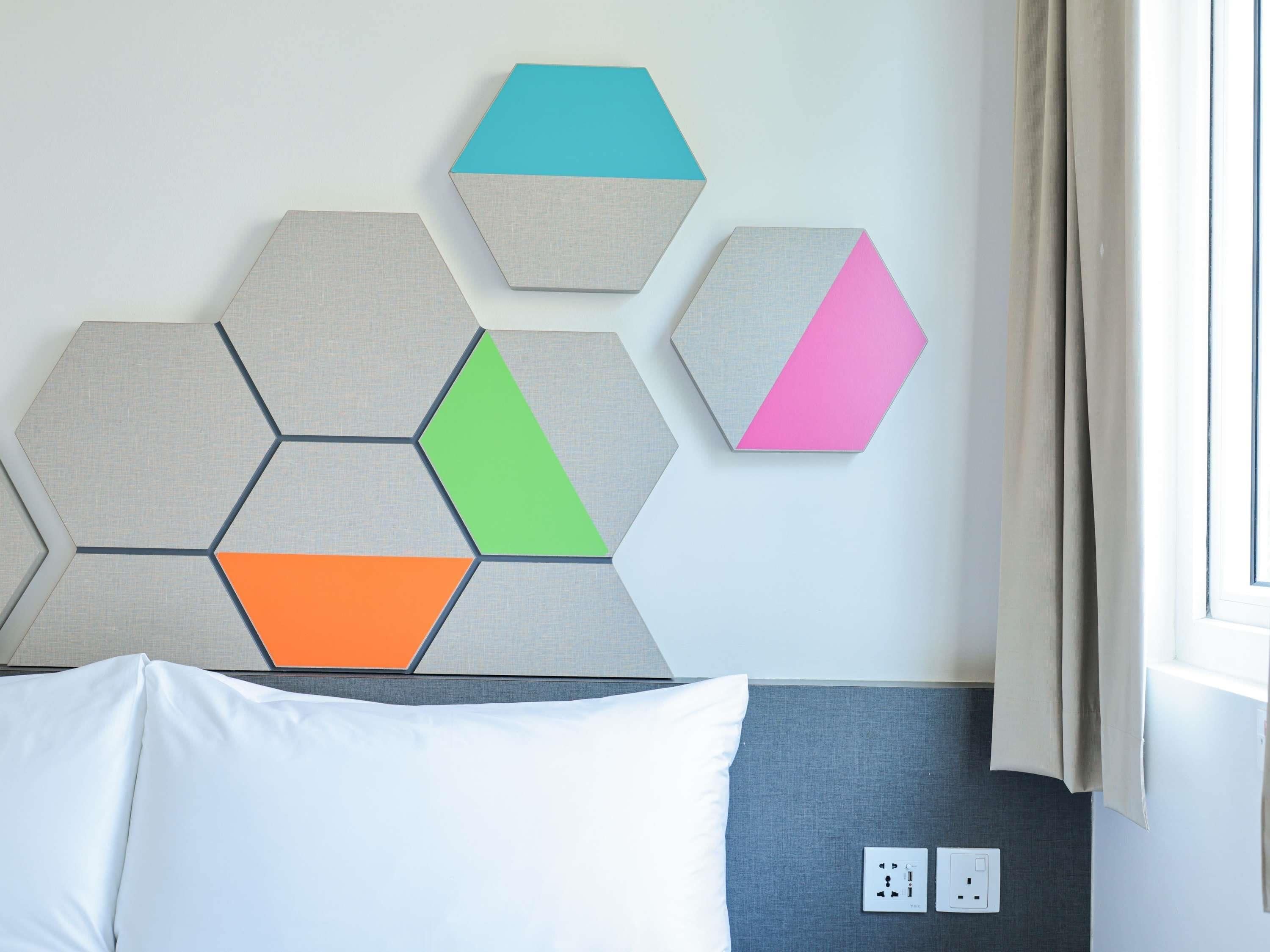 Une chambre d’hôtel comprenant un lit, une œuvre d’art géométrique accrochée au mur, une fenêtre avec des rideaux et des prises électriques.