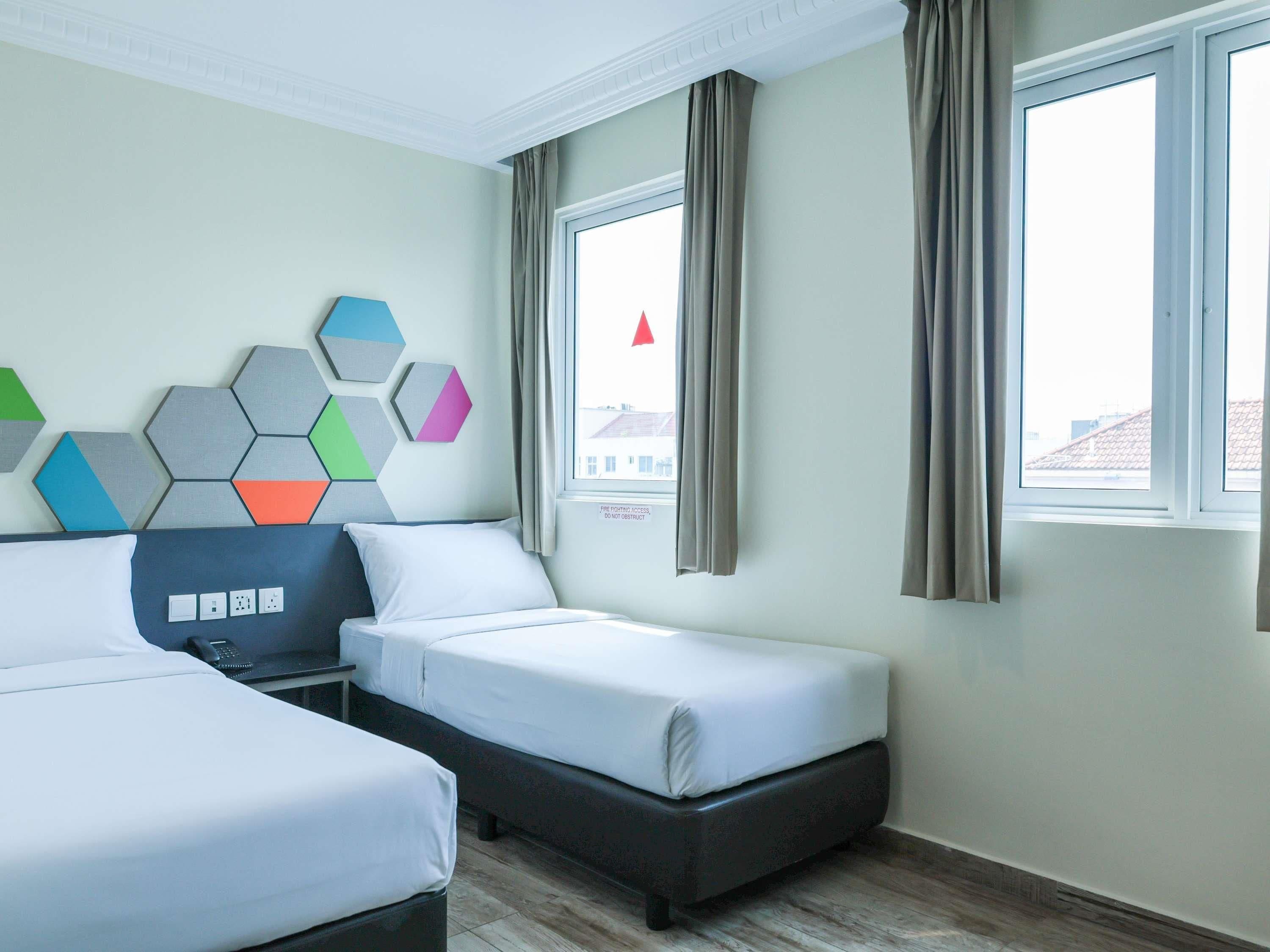 Una habitación de hotel con dos camas, una mesita de noche, una lámpara de pared y un ventanal con cortinas.