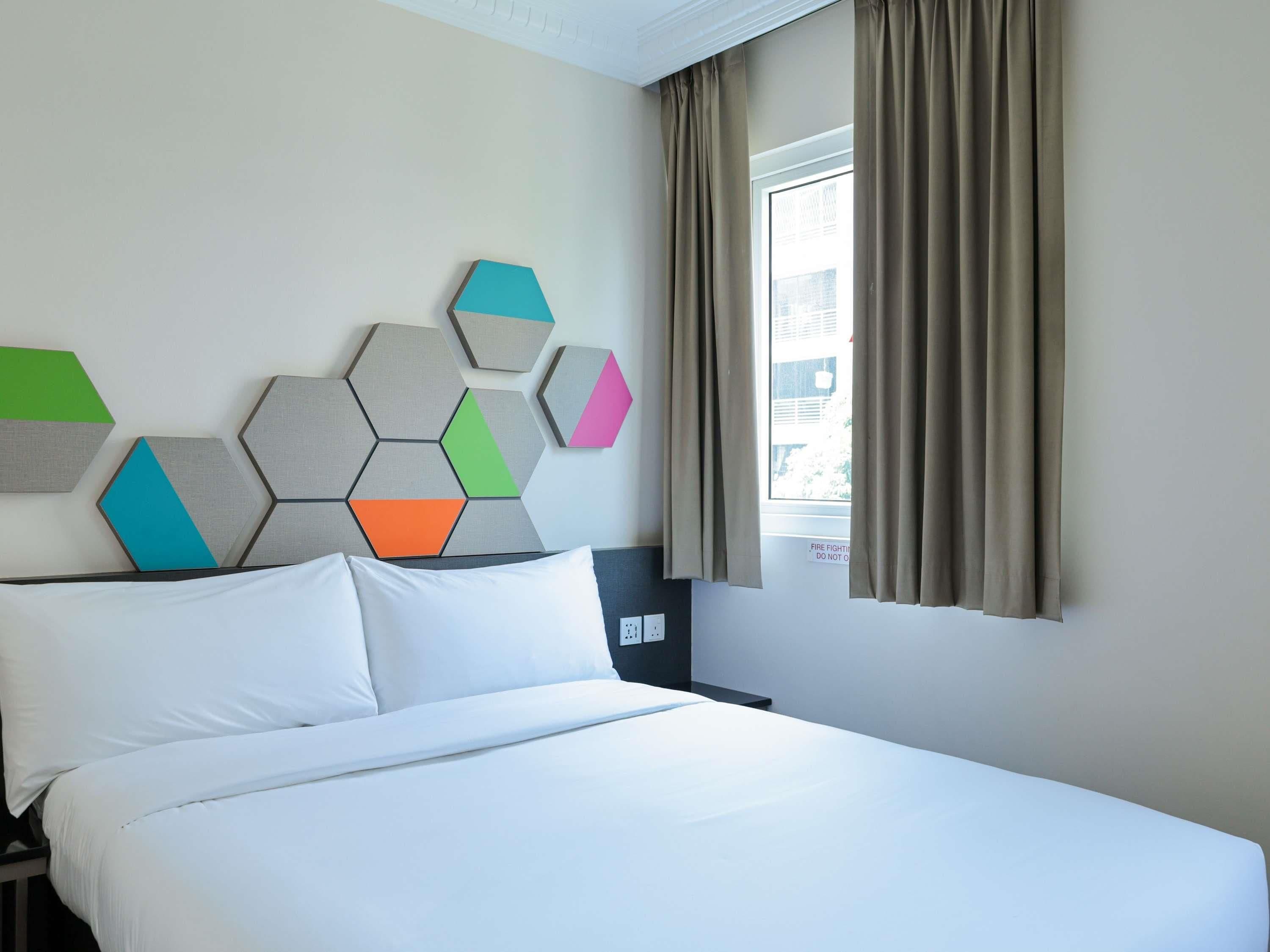 A hotel room with a bed, a wall-mounted geometric art piece, a window with curtains, and a nightstand.
