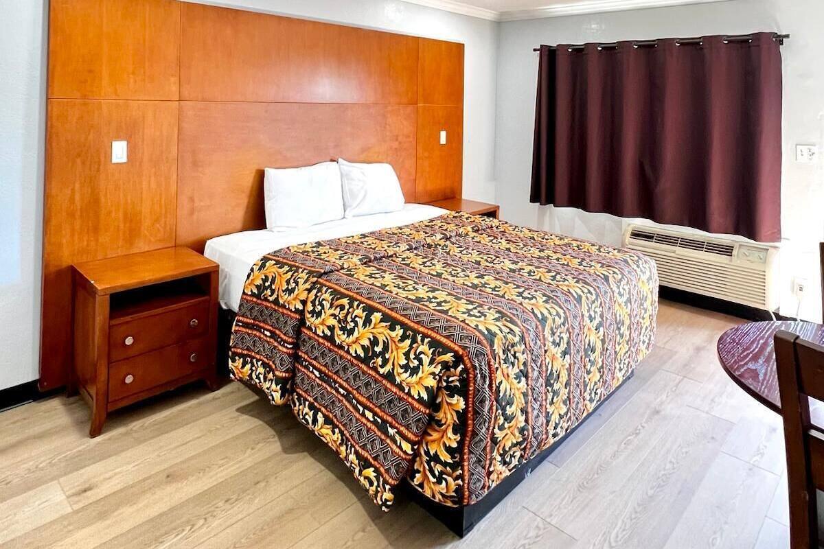 A bedroom with a wooden headboard, a bed with a patterned bedspread, a nightstand, a chair, and a window with curtains.
