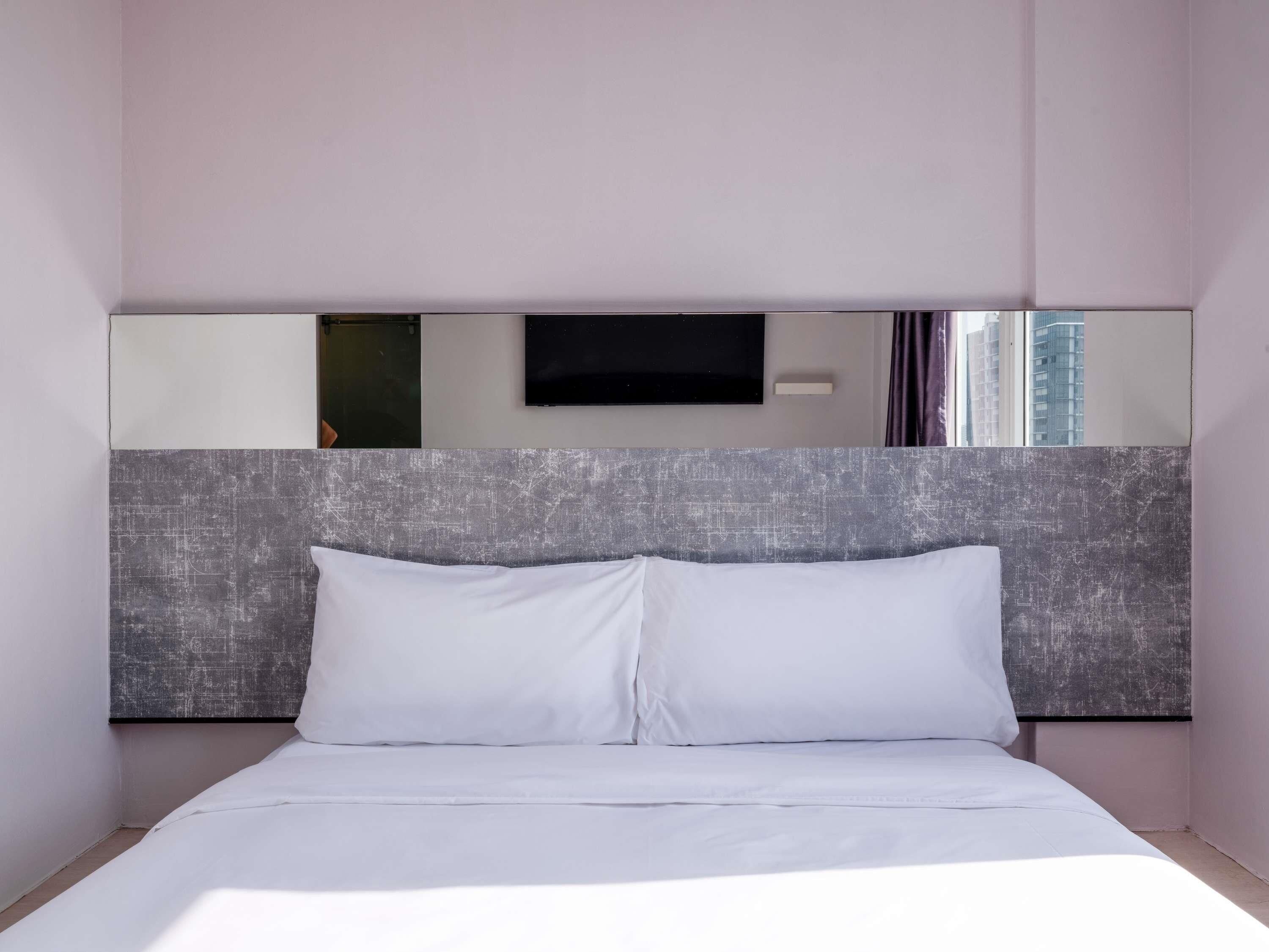 A modern hotel room with a large bed, a headboard with a textured design, a flat-screen TV mounted on the wall, and a window with a view of buildings.