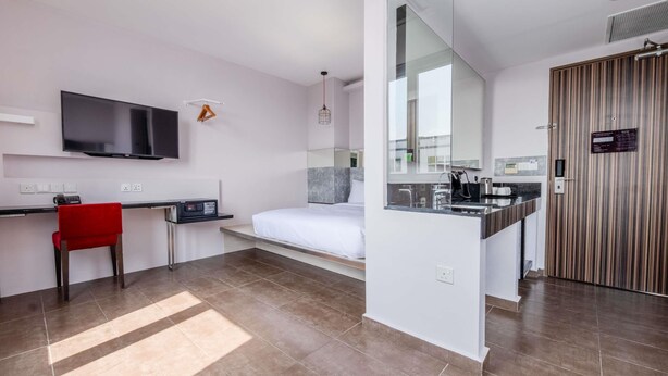 Una habitación de hotel moderna con una cama, un escritorio, un televisor, una silla roja y un baño con espejo y lavamanos.