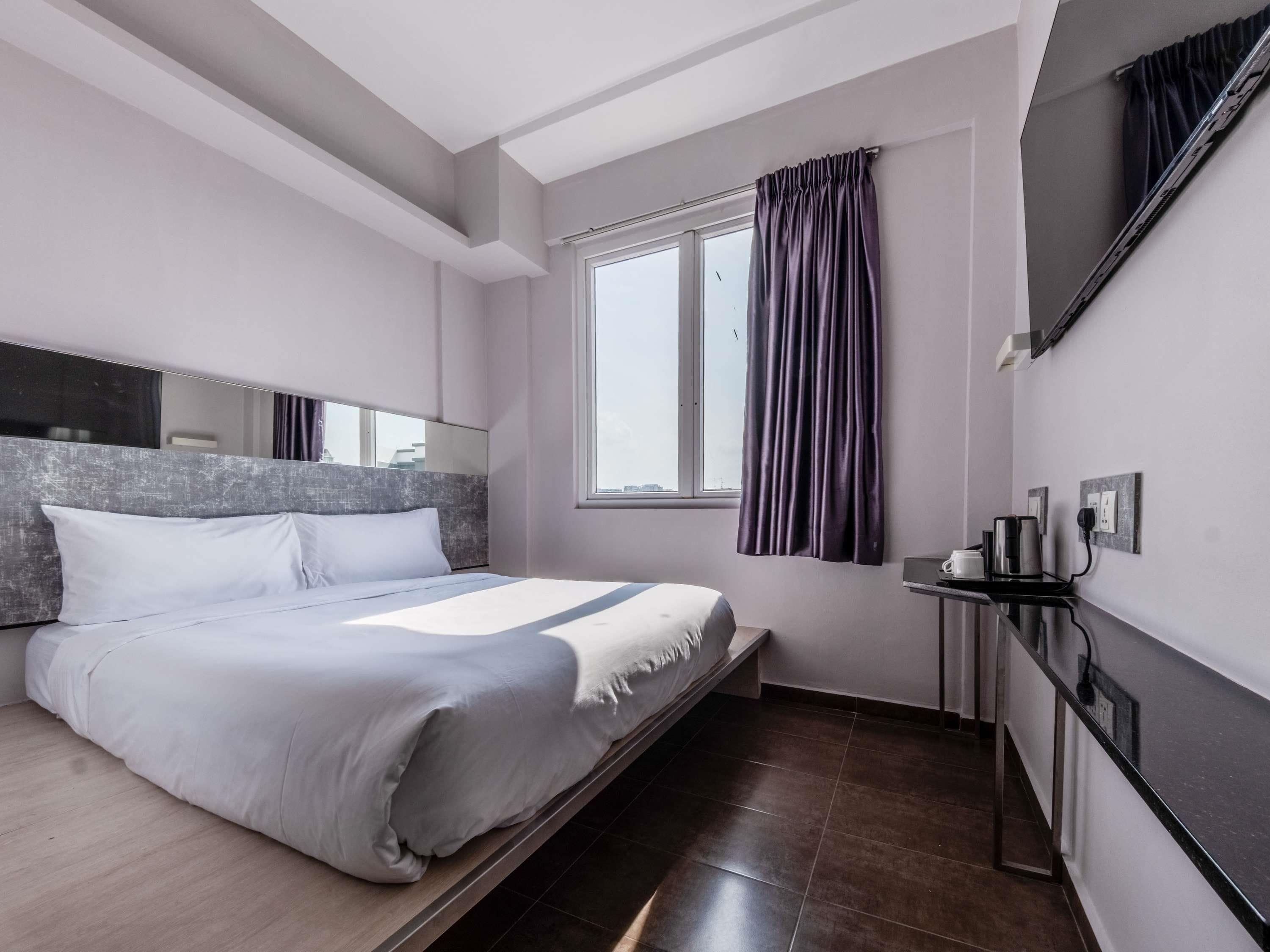 A modern hotel room with a large bed, a bedside table with a flat-screen TV, a window with curtains, and a view of the city.