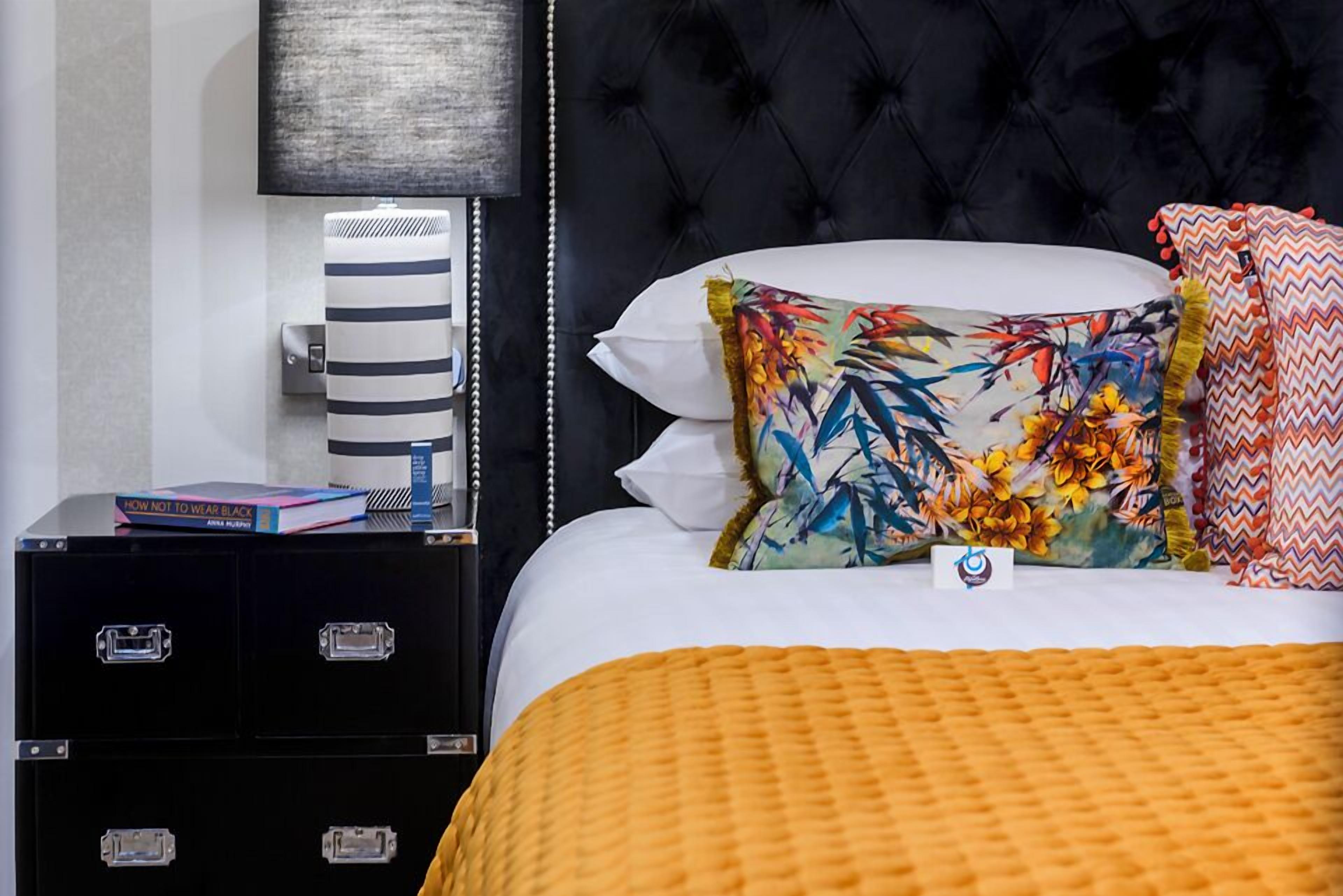 A neatly made bed with a white pillow and a colorful floral pillow, a black nightstand with a striped lamp, and a black storage unit with drawers.
