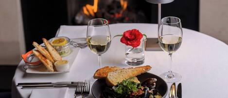 A dining table with a bowl of mussels, two glasses of white wine, a red rose, bread, and a candle.