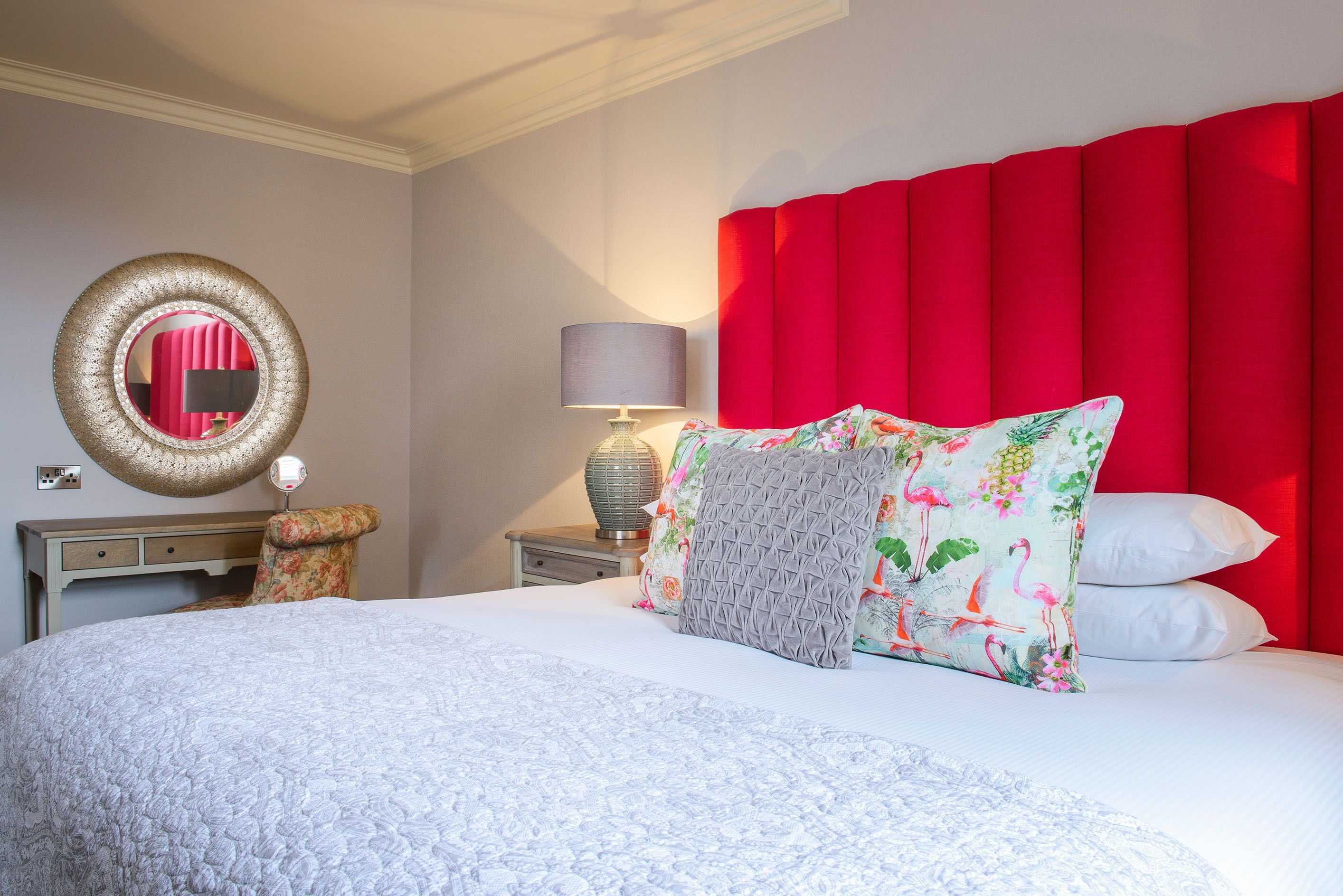 Una cama bien hecha con sábanas blancas y varios almohadones, un espejo redondo en la pared, una lámpara sobre una mesita de noche y un cabecero rojo.