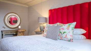 Una cama bien hecha con sábanas blancas y varios almohadones, un espejo redondo en la pared, una lámpara sobre una mesita de noche y un cabecero rojo.