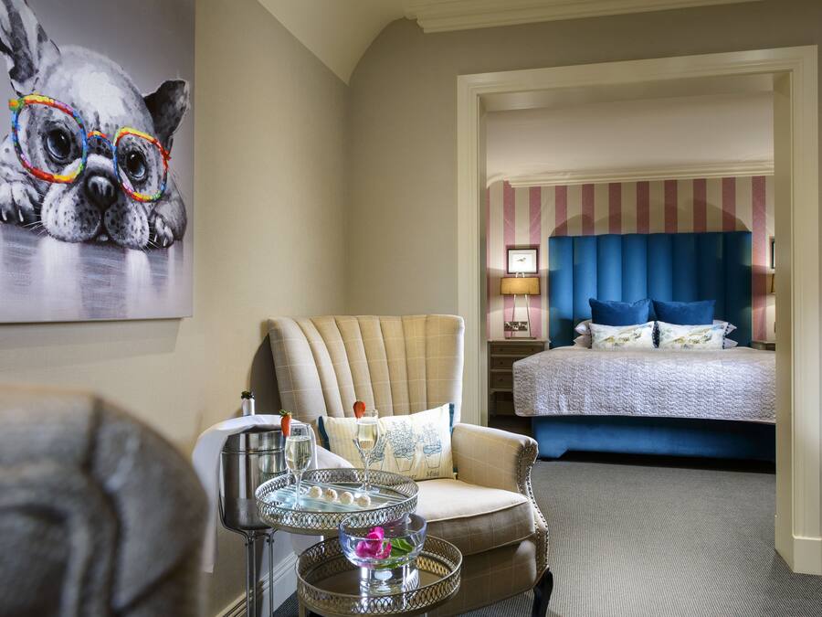 A hotel room with a bed, a chair, a table with a drink, and a painting on the wall.
