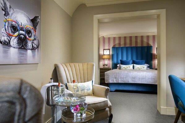 A hotel room with a bed, a chair, a table with a drink, and a painting on the wall.