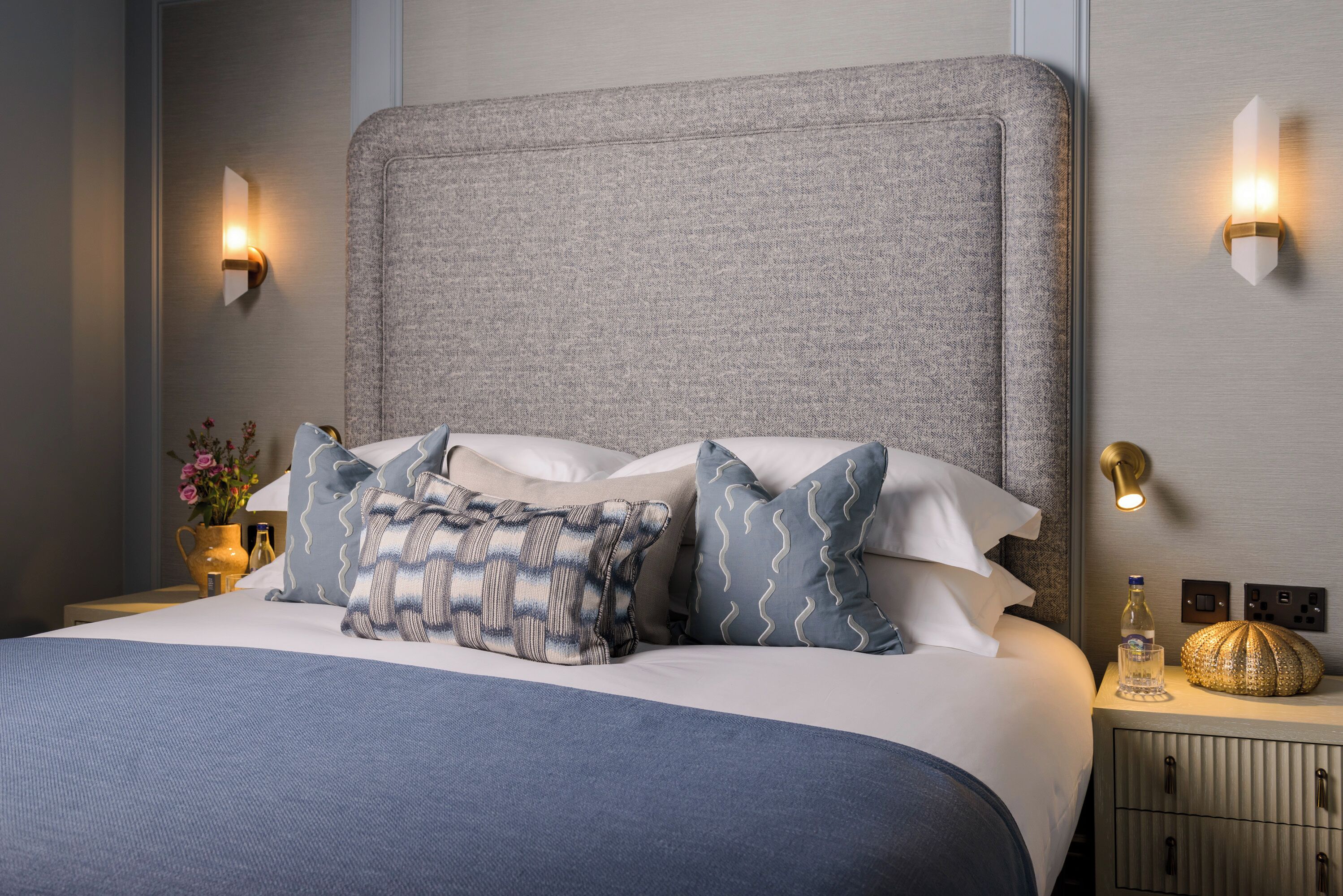 A neatly made bed with a grey headboard, multiple pillows, bedside lamps, and a small bedside table with a bottle and a decorative item.