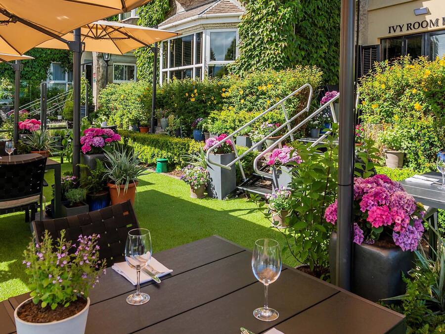Un patio al aire libre con mesas y sillas, rodeado de plantas y flores en macetas.