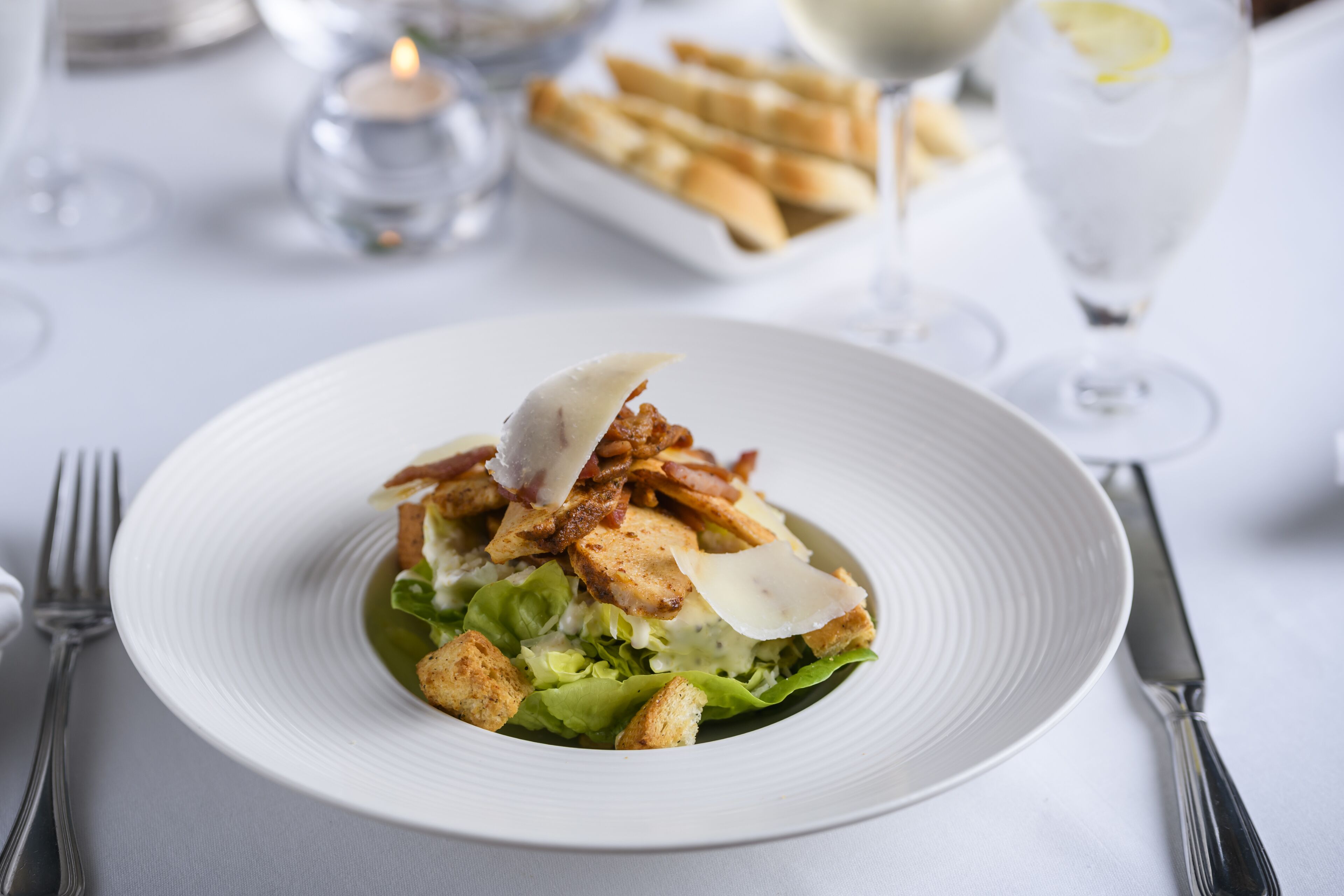 Een witte schaal met een salade, croutons en spek op een witte tafelkleed.