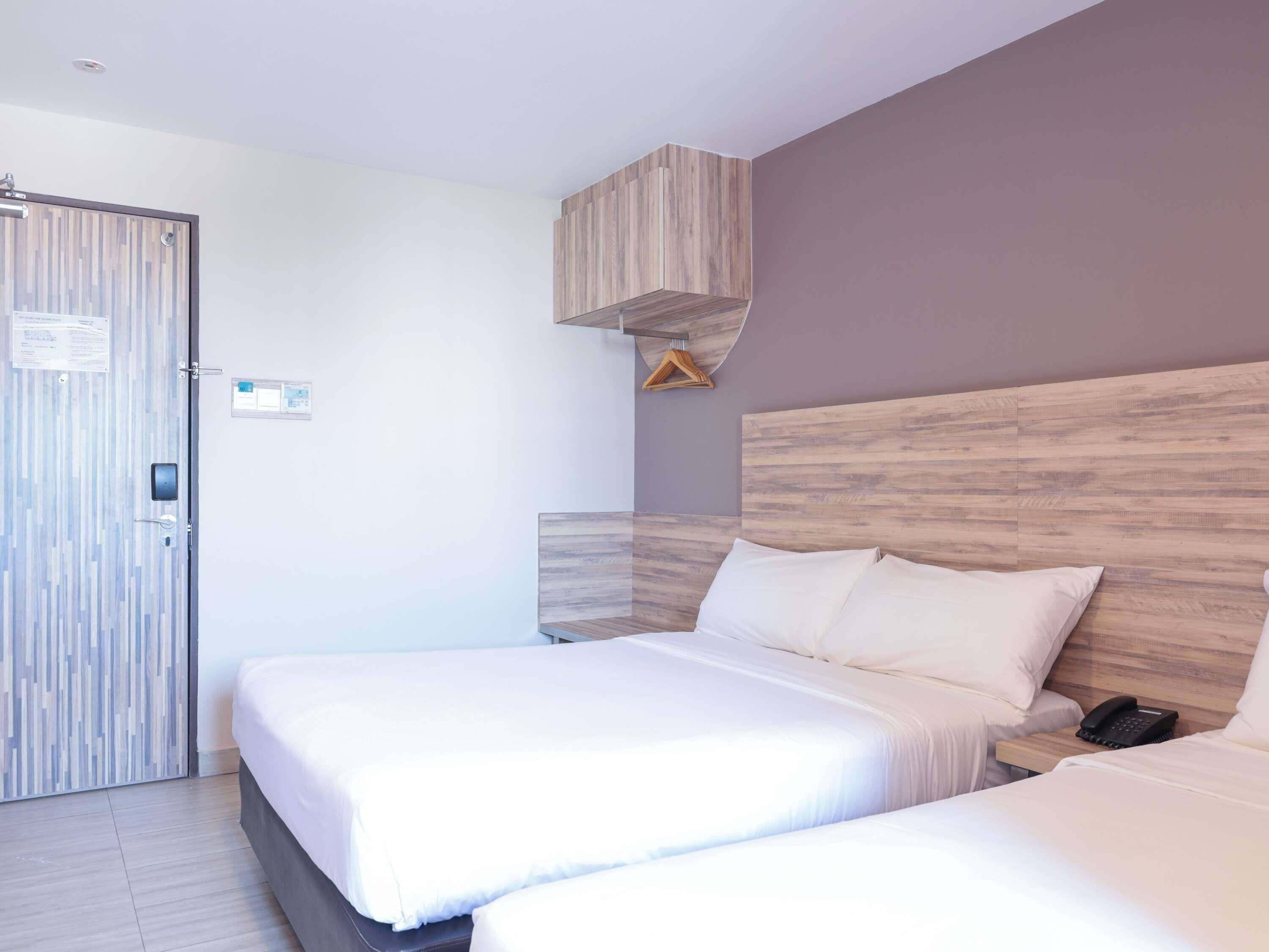 A hotel room with two beds, a wooden headboard, a bedside table with a phone, and a door with a lock and a notice.