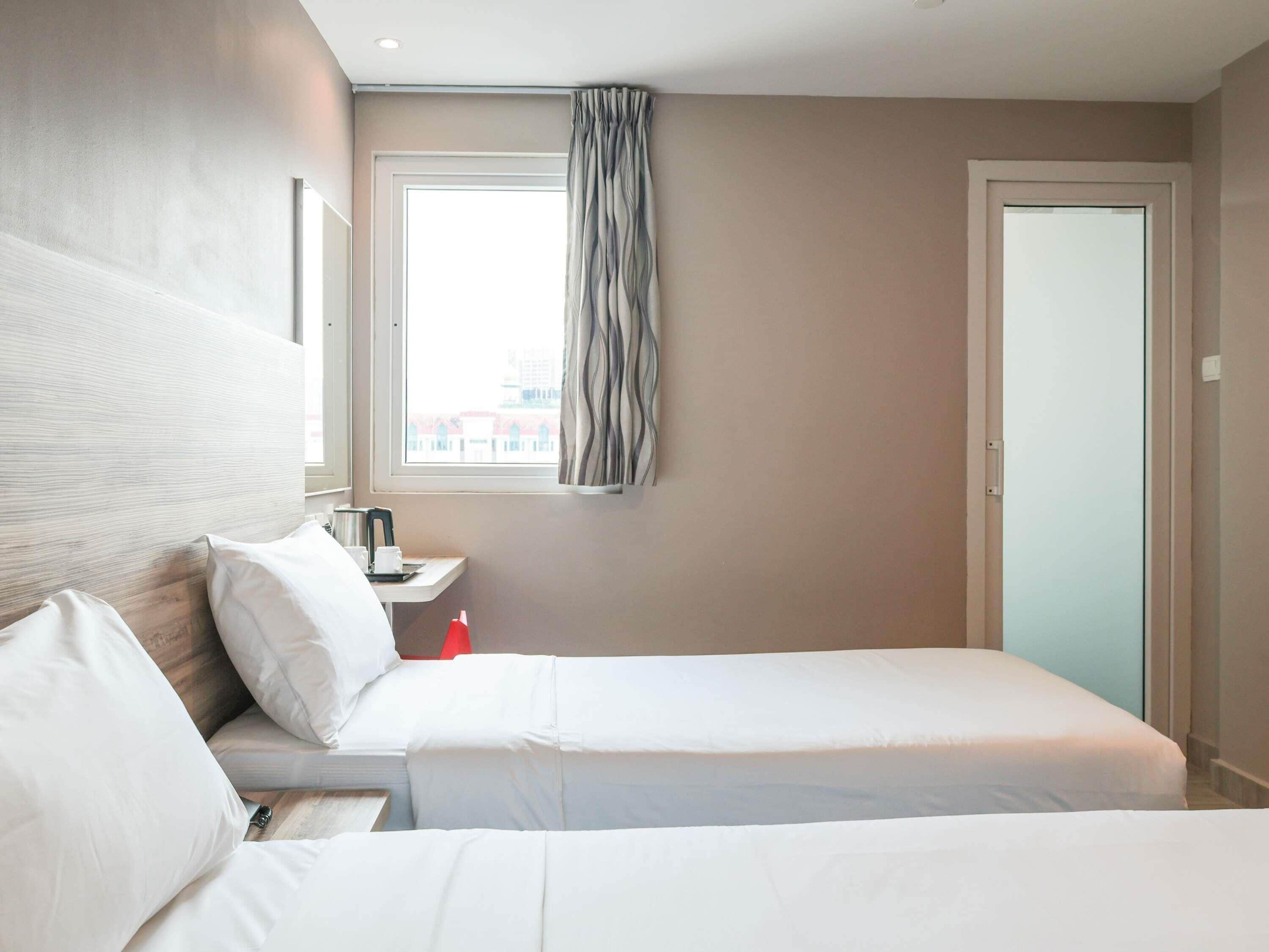 A hotel room with two beds, a window with curtains, a bedside table with a kettle, and a door.