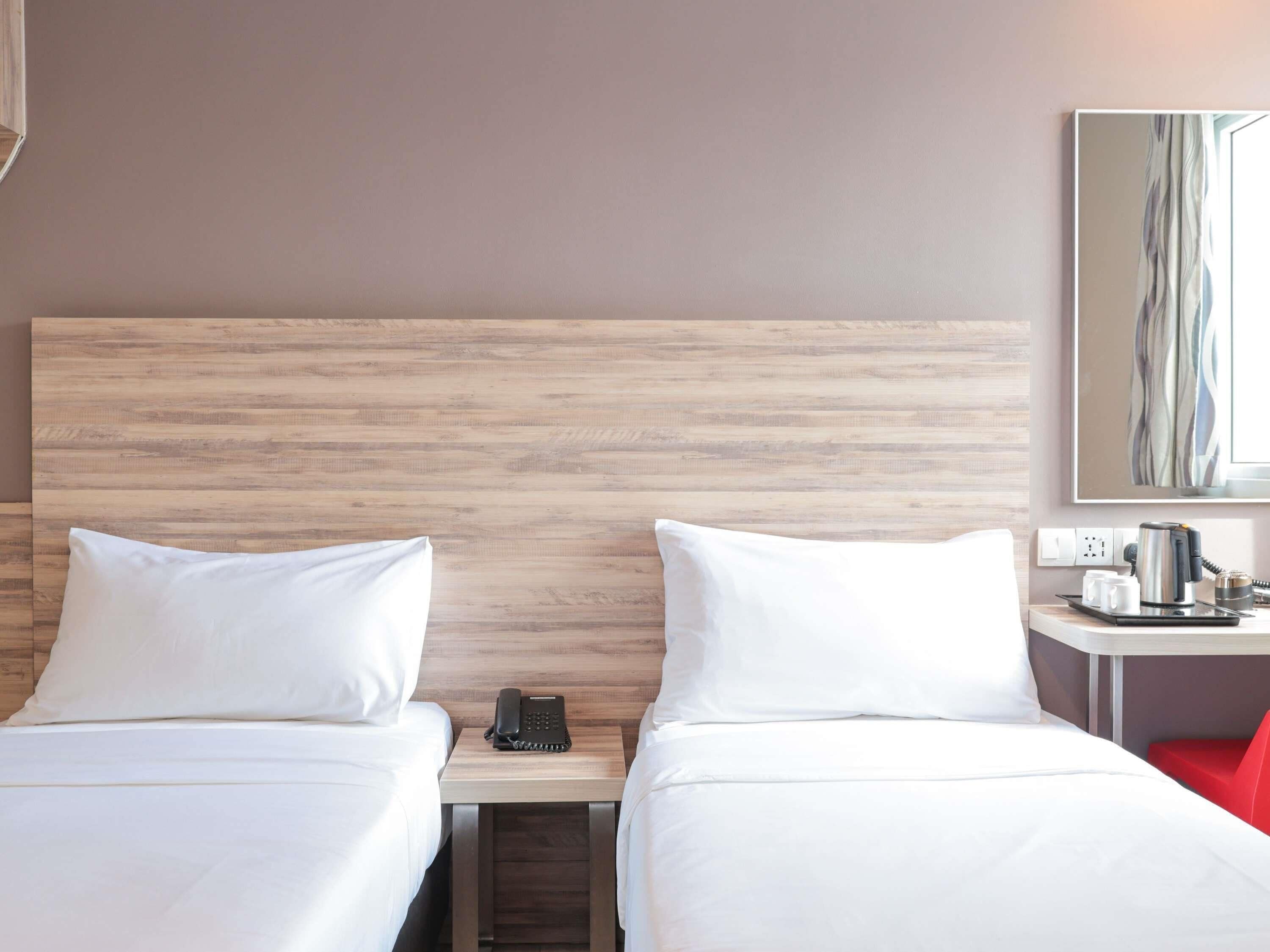 A hotel room with two beds, a wooden headboard, a bedside table with a telephone, a red chair, and a wall-mounted artwork.