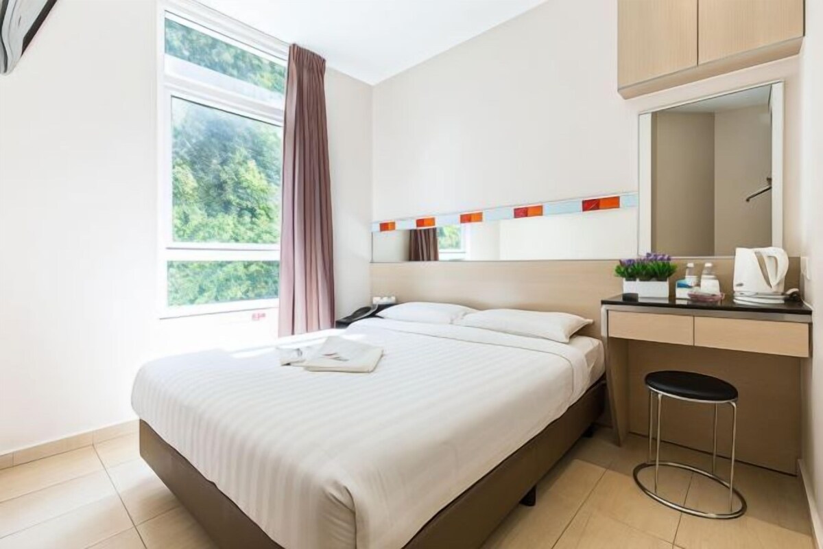 A modern hotel room with a large bed, a desk with a chair, a mirror, and a window with curtains.