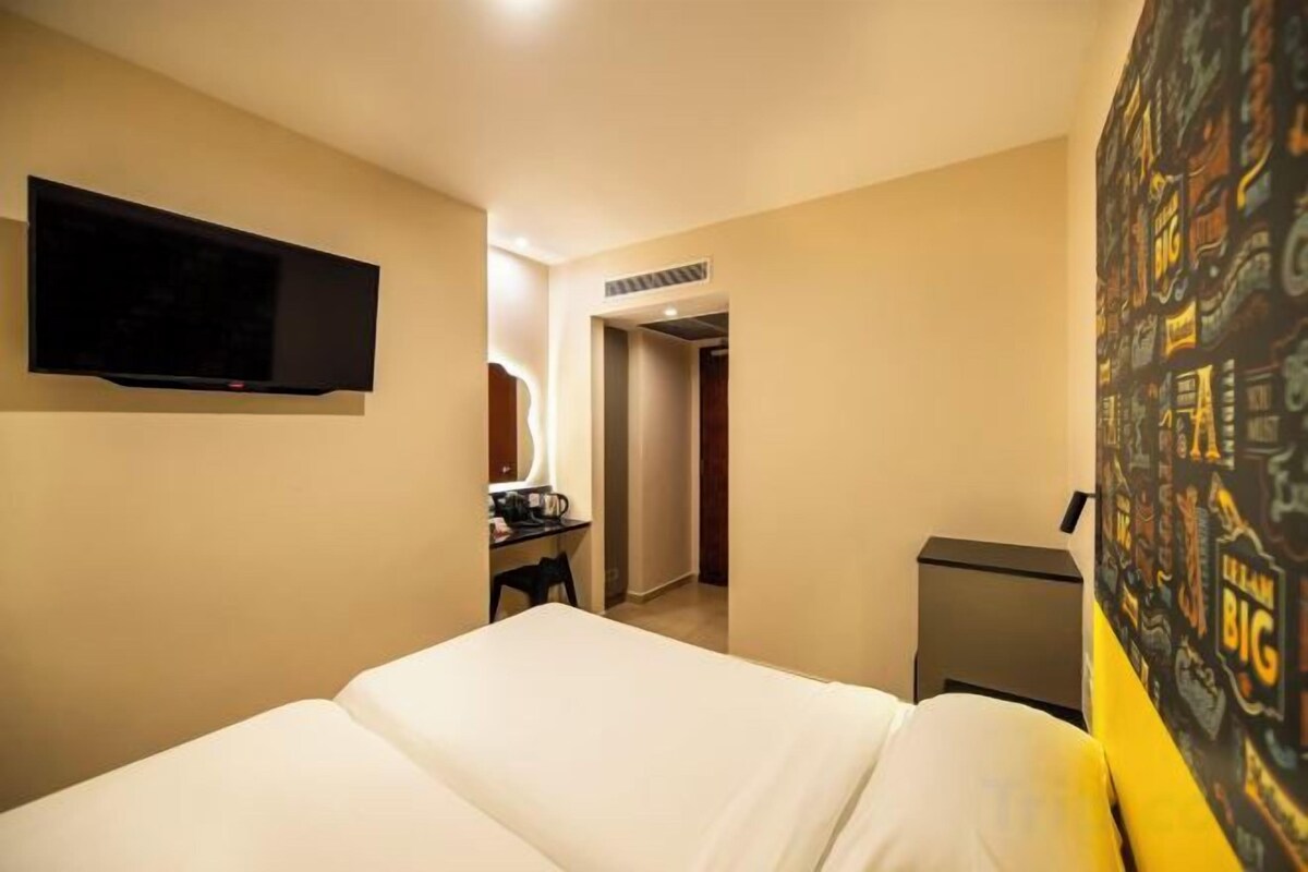 A hotel room with a bed, a desk with a chair, a television mounted on the wall, and a door leading to another room.