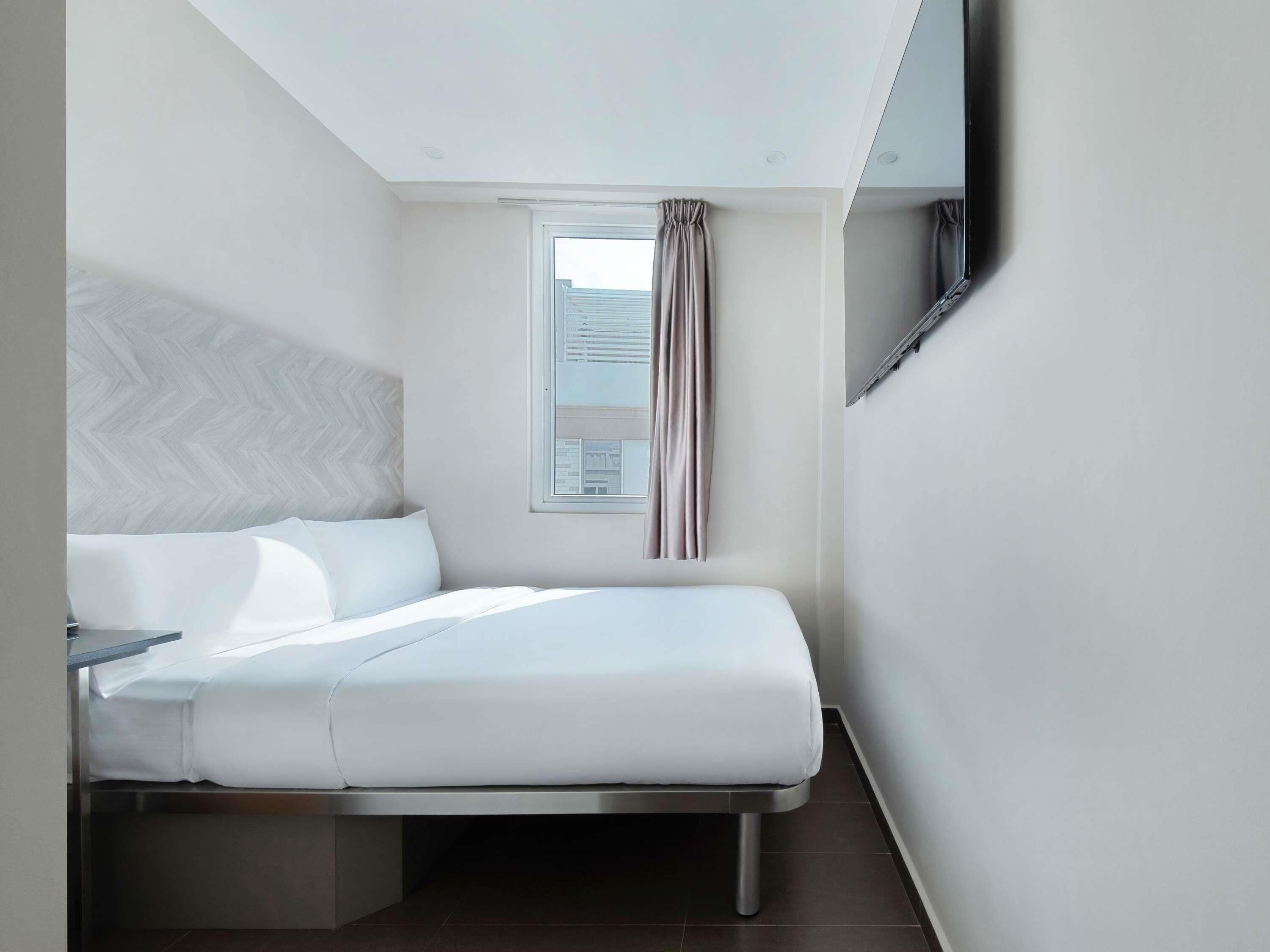 A modern hotel room with a bed, a window with curtains, a flat-screen TV mounted on the wall, and a herringbone-patterned headboard.