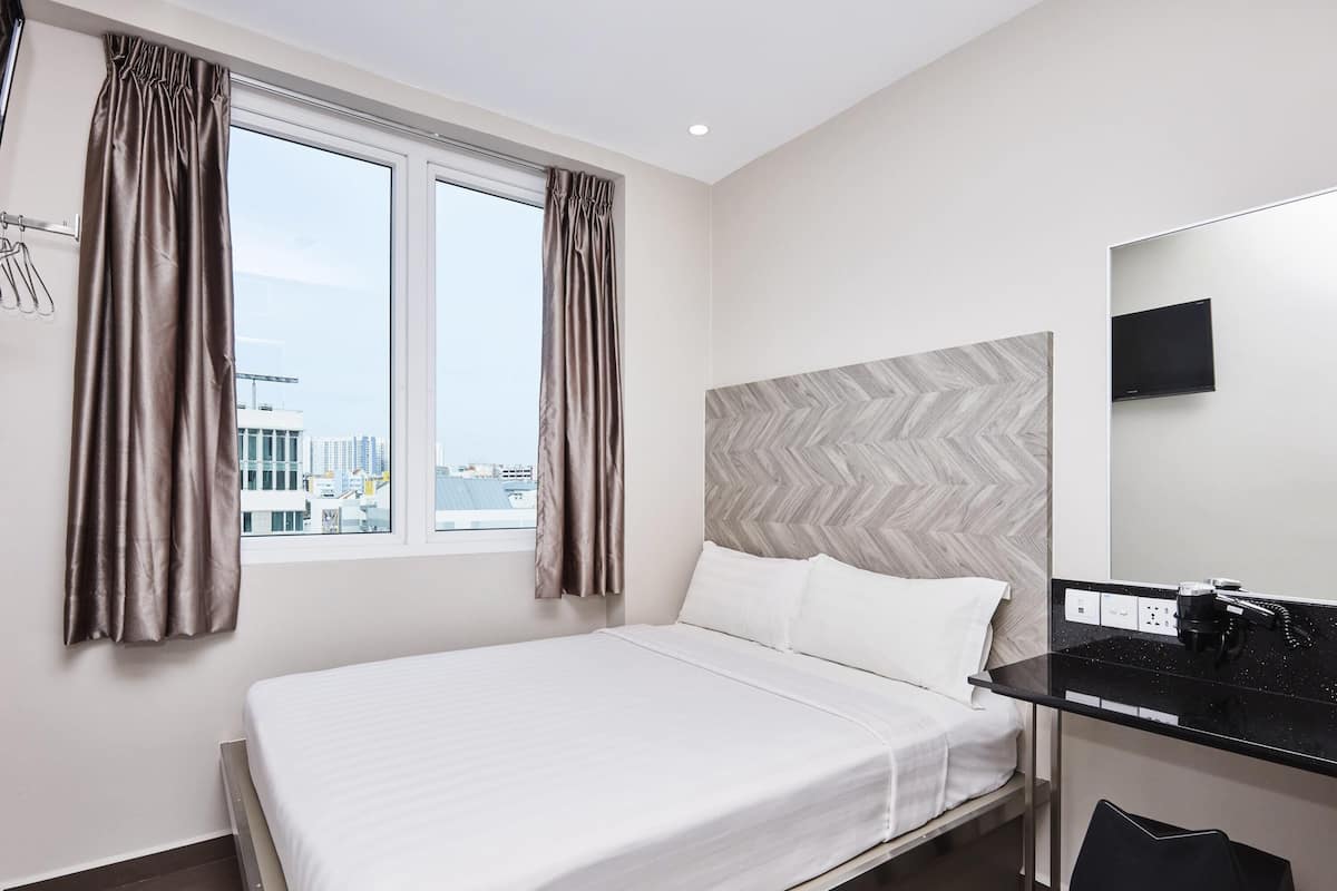 A hotel room with a bed, a desk with a mirror, a chair, and a view of the cityscape through the window.
