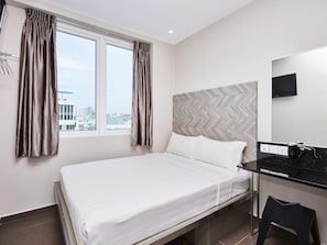 A hotel room with a bed, a desk with a mirror, a chair, and a view of the cityscape through the window. - ibis budget Singapore Ruby (Singapore)