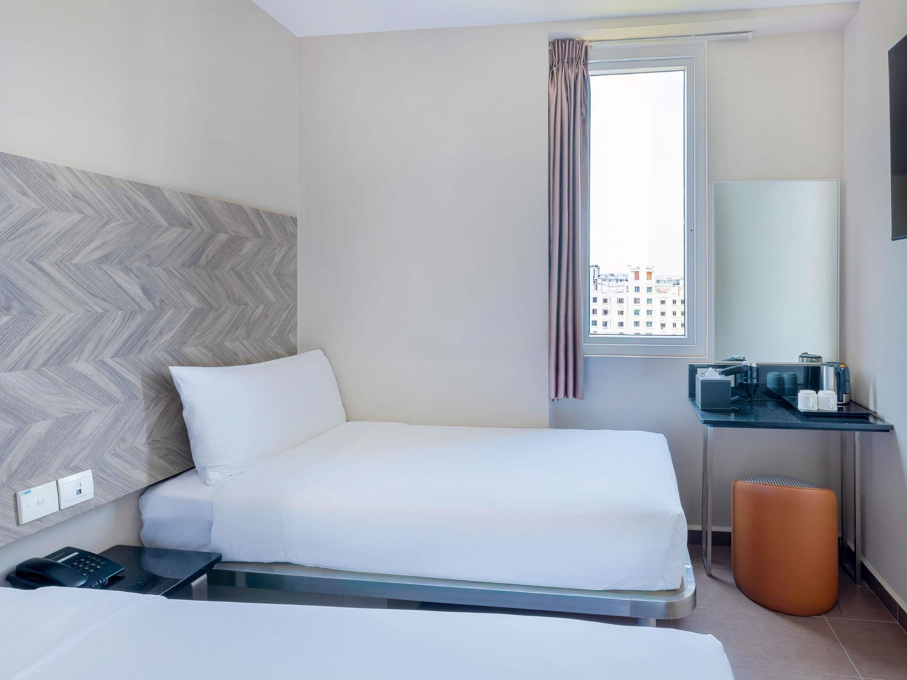 Habitación de hotel con cama, escritorio con espejo, ventana con cortinas y vista a la ciudad.
