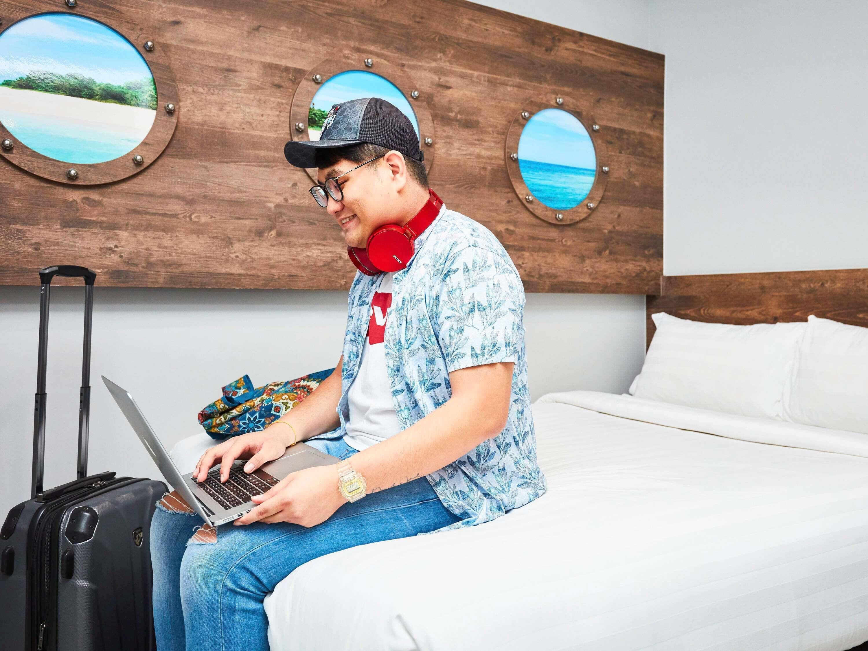 Une personne assise sur un lit, avec un ordinateur portable, une valise et un mur orné de fenêtres rondes.