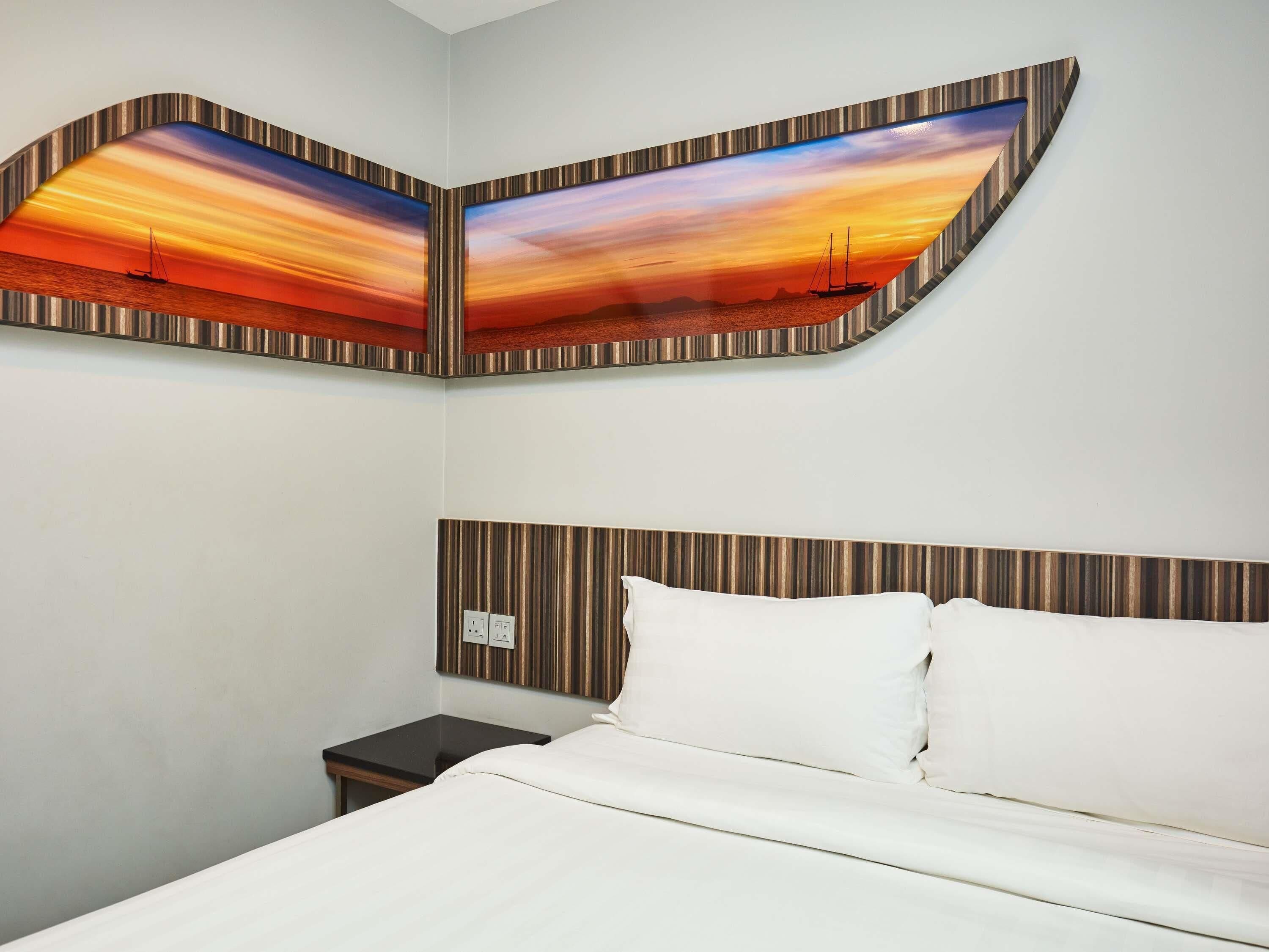 A hotel room with a bed, a nightstand, and a wall-mounted painting of a sunset over the ocean.