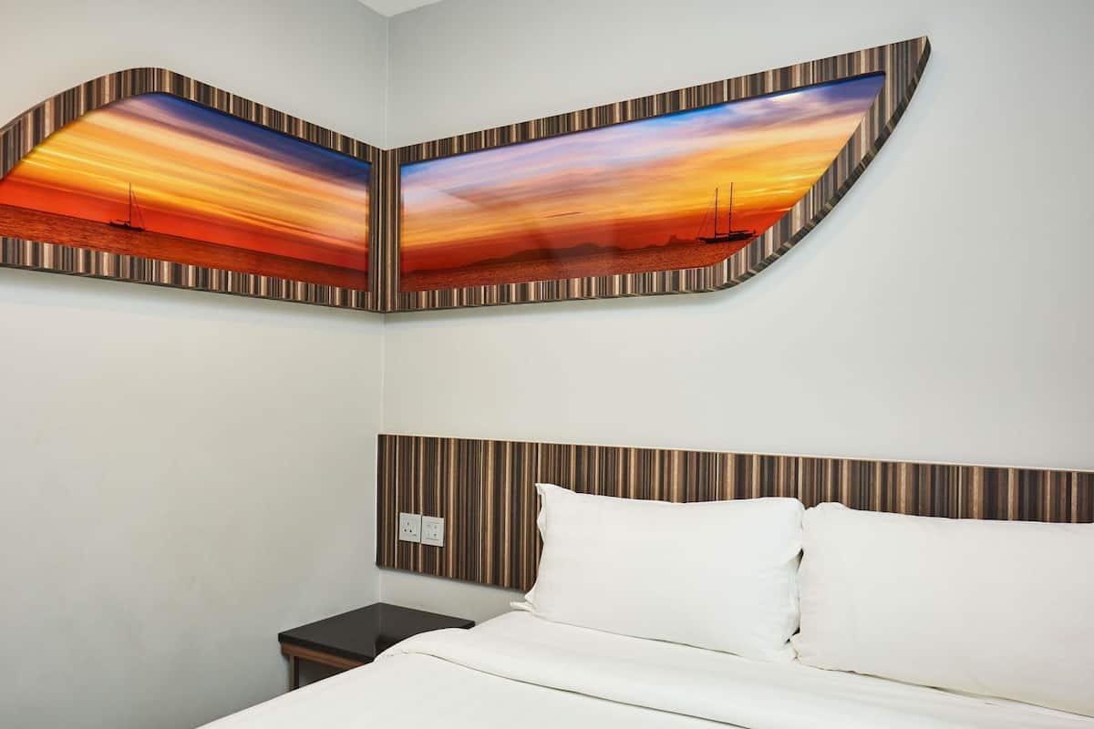 A hotel room with a bed, a nightstand, and a wall-mounted painting of a sunset over the ocean.