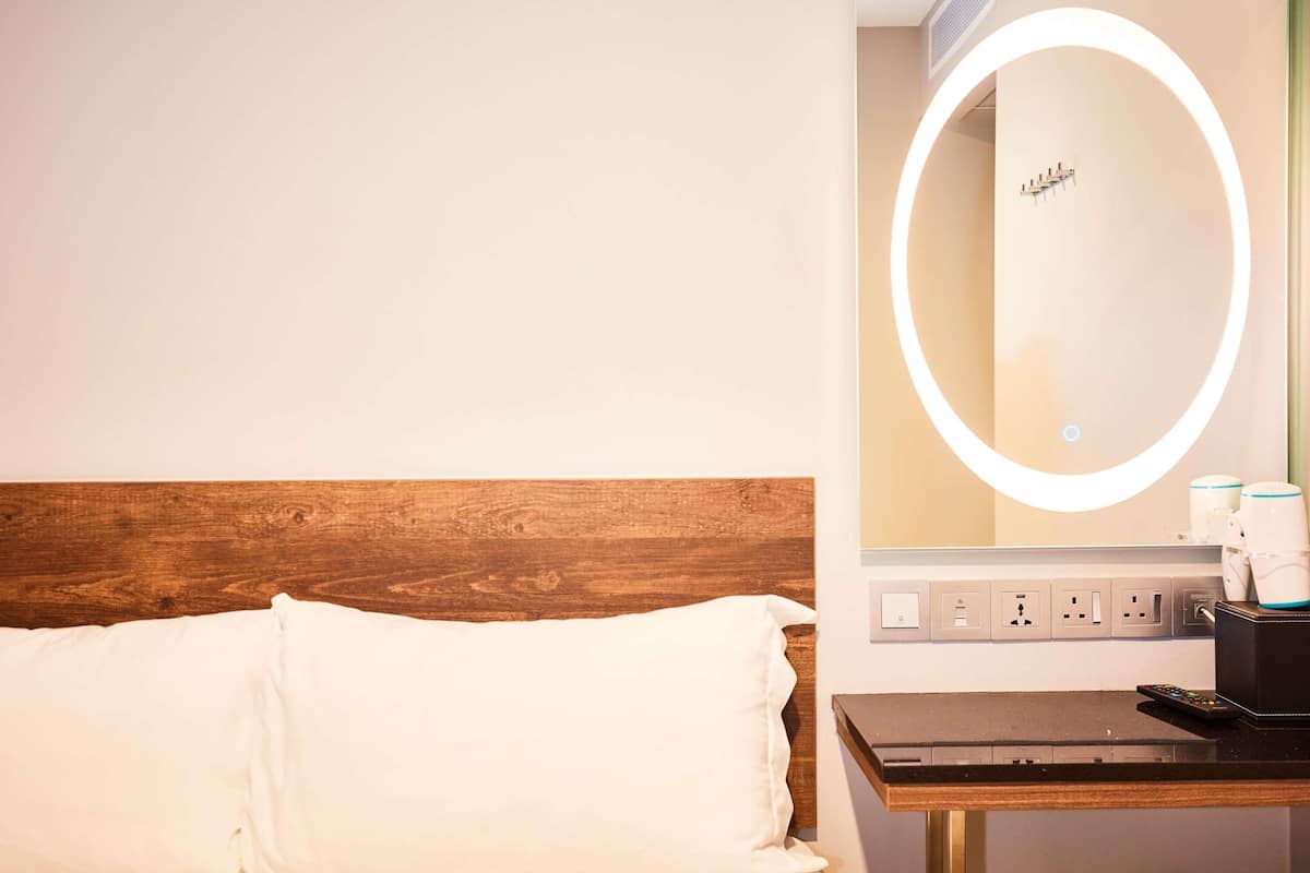 A hotel room with a wooden headboard, a bed with white bedding, a bedside table with a remote control, and a wall-mounted mirror with a light.