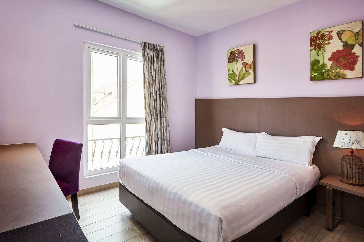 A hotel room with a bed, a chair, a desk, and a window with curtains.