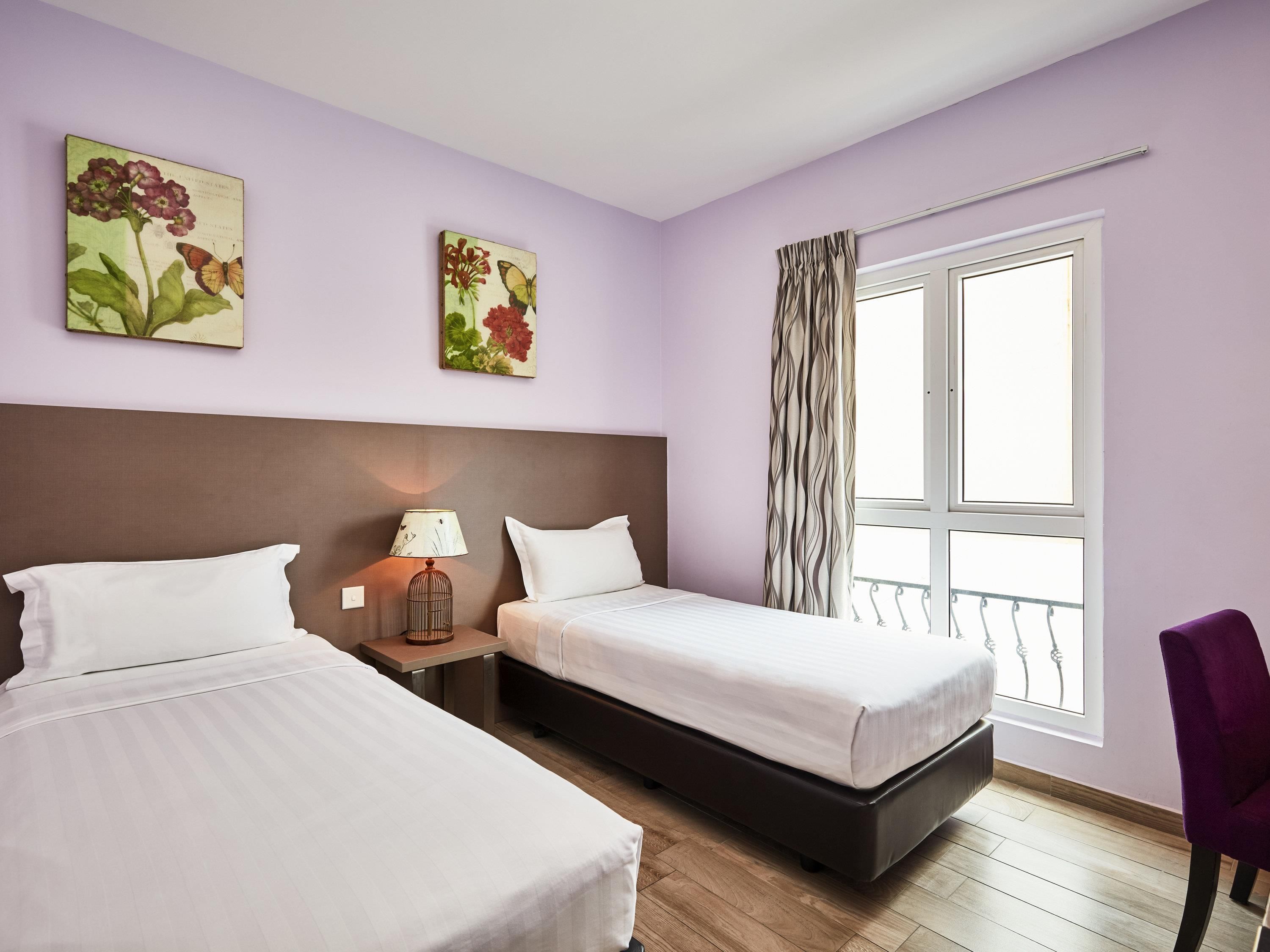 A hotel room with two beds, a nightstand with a lamp, and a window with curtains.