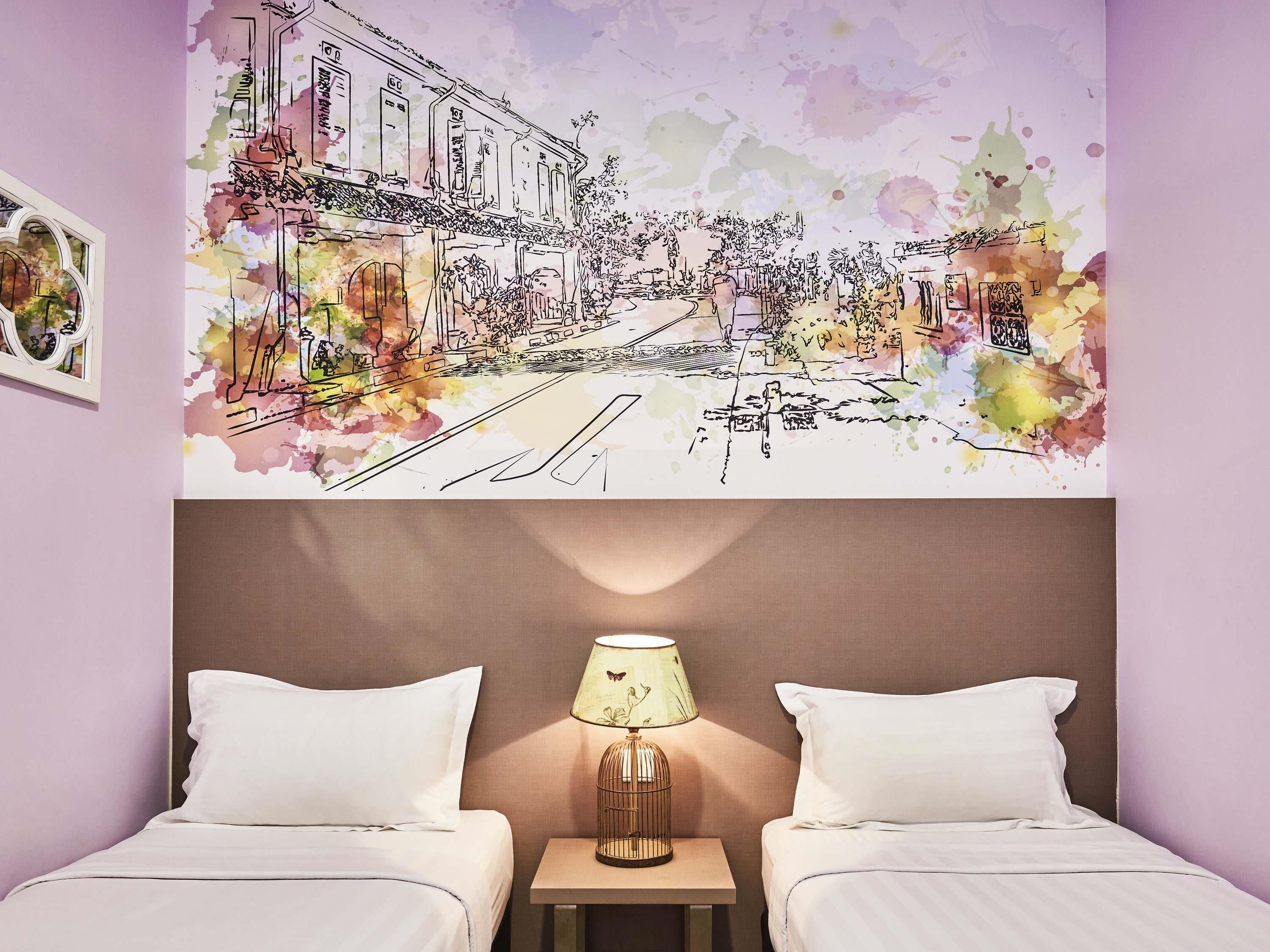 A hotel room with a bed, two pillows, a nightstand with a lamp, and a wall mural depicting a street scene.