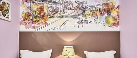 Habitación de hotel con una cama, dos almohadas, una mesita de noche con lámpara y un mural en la pared que muestra una escena callejera.
