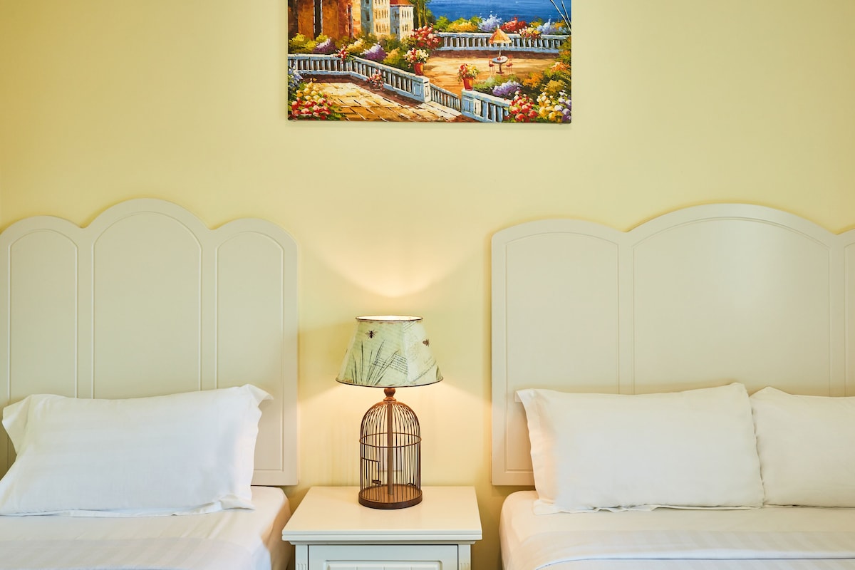 A hotel room with two beds, a nightstand with a lamp, and a painting of a coastal scene on the wall.