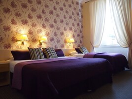 A hotel room with two beds, a floral wallpaper, two wall-mounted lamps, and a window with curtains.