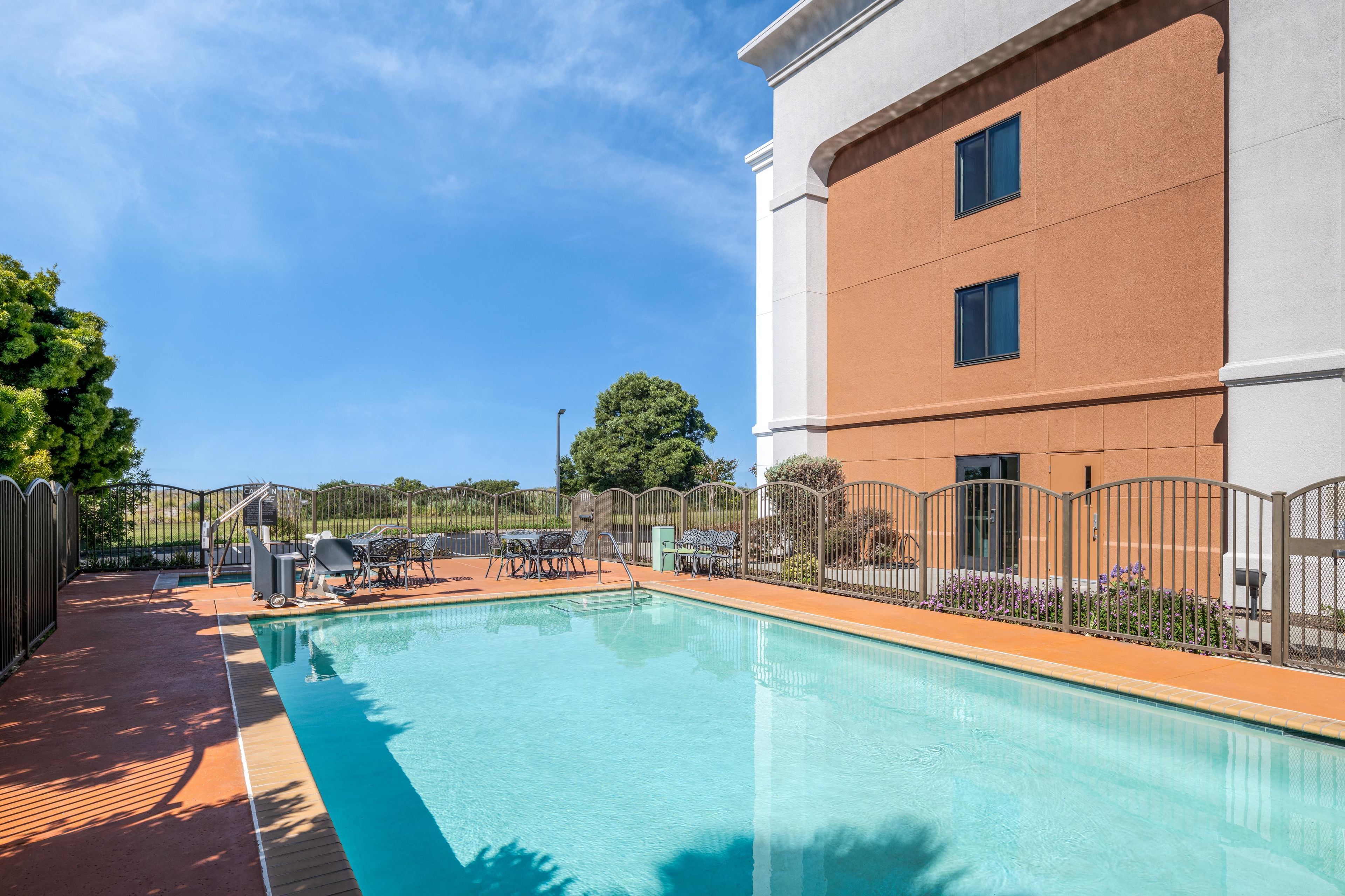 Une piscine avec un espace patio, entourée d’une clôture et bordée d’un bâtiment doté de deux fenêtres.