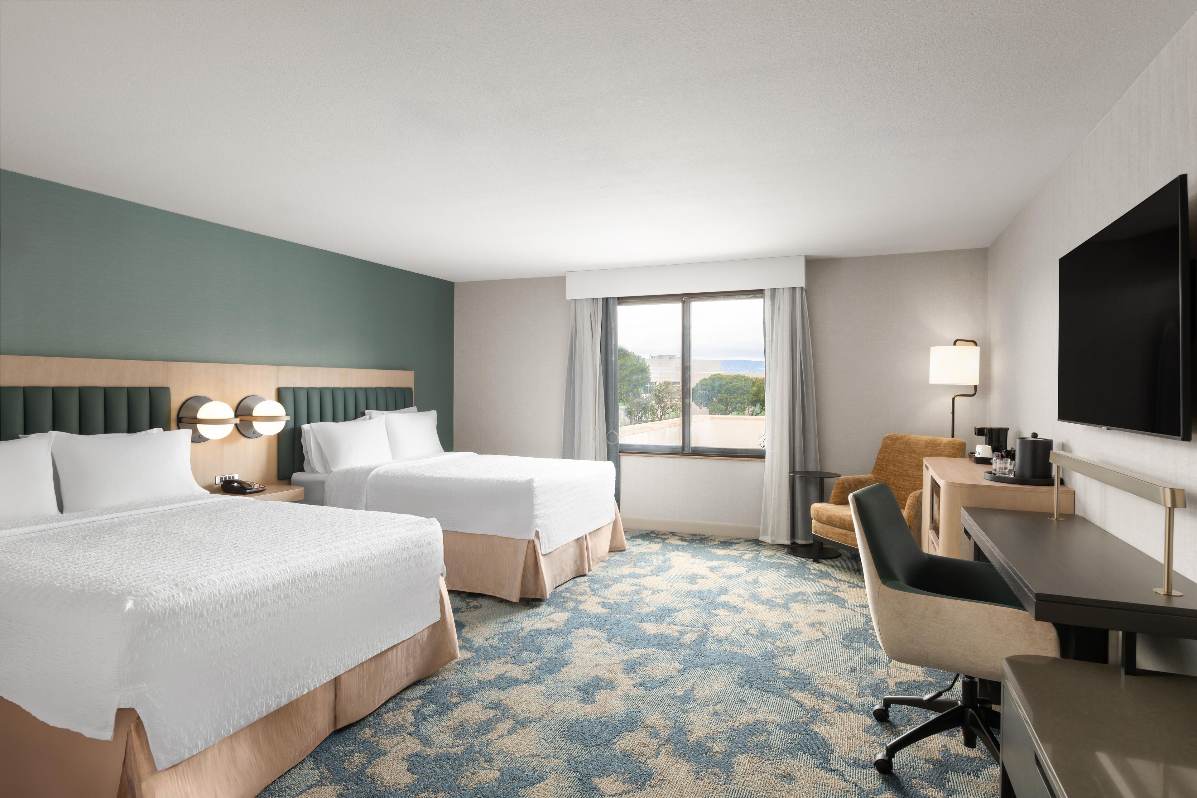 Habitación de hotel con dos camas, un escritorio, una silla, un televisor y una ventana con vistas.