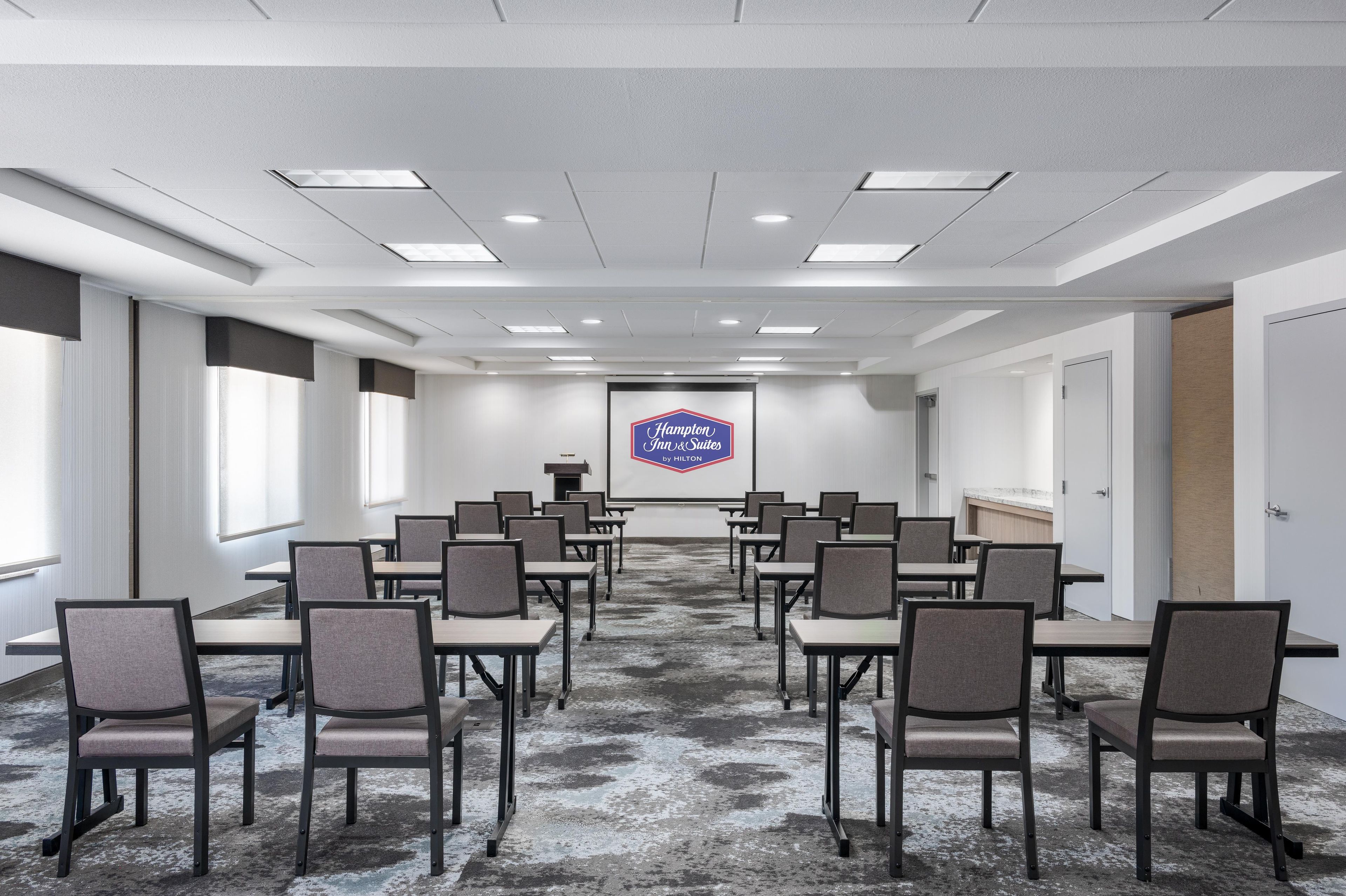 A conference room with a large screen displaying the Hampton Inn logo, rows of chairs, and a carpeted floor.