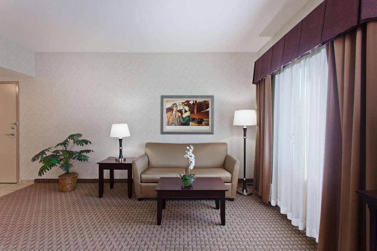 Een hotelkamer met een beige bank, twee bijzettafels met lampen, een ingelijst schilderij aan de muur, een potplant en een groot raam met gordijnen.