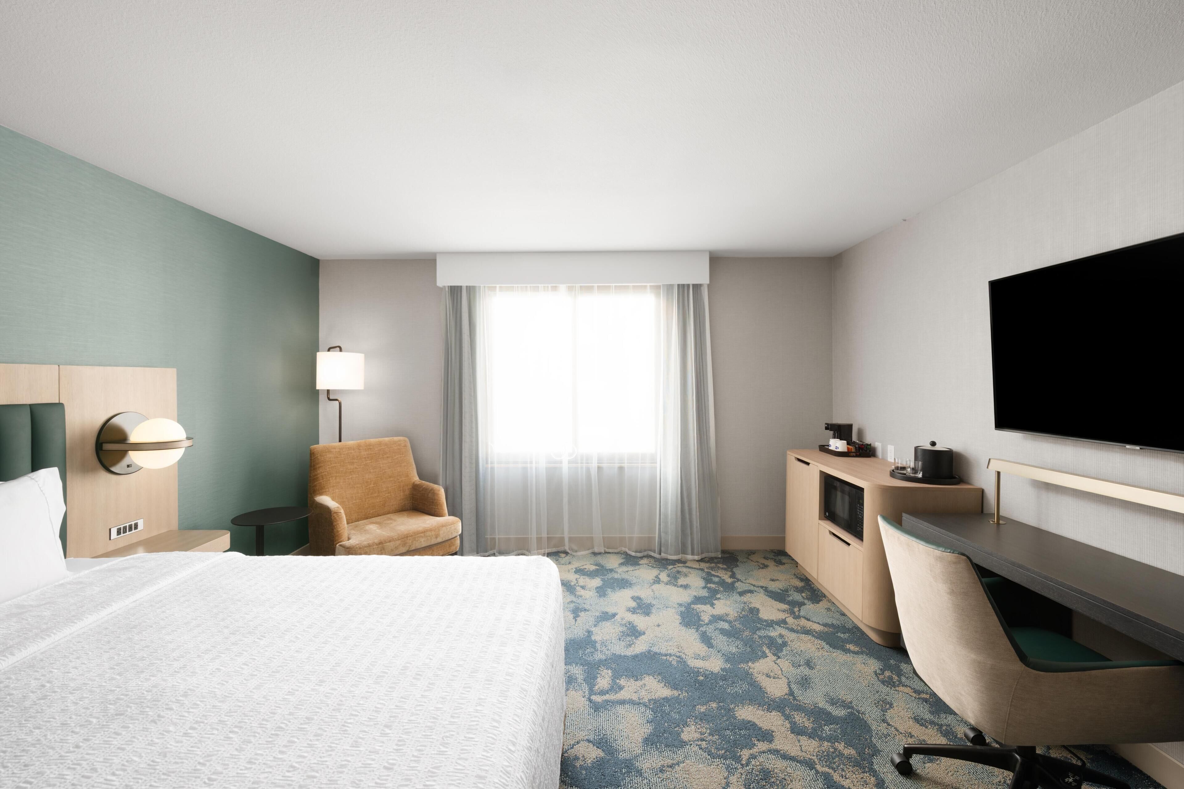A hotel room with a bed, a desk, a chair, a TV, and a window with curtains.