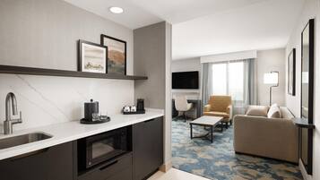 A modern hotel room with a kitchen area, a living space with a sofa and armchair, and a flat-screen TV mounted on the wall.