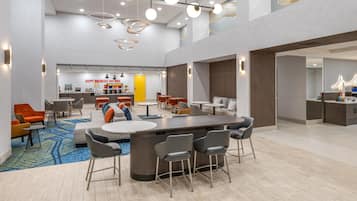 Un hall d’hôtel moderne avec un bar central, des espaces aménagés pour s’asseoir et une cuisine en arrière-plan.