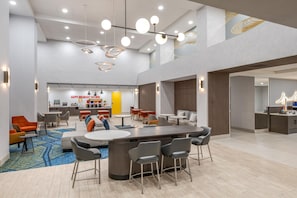 A modern hotel lobby with a central bar area, seating arrangements, and a kitchen in the background.