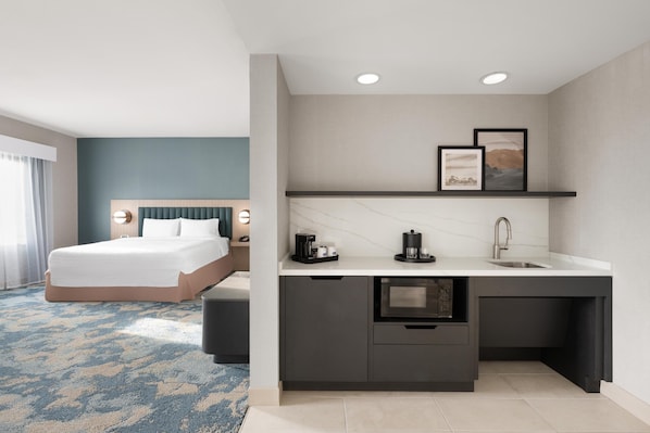 A modern hotel room with a bed, a small kitchenette, and a bathroom.